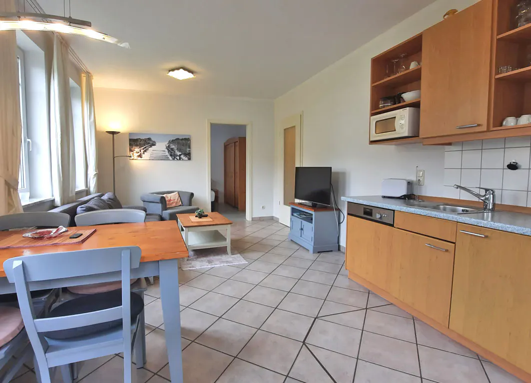 Open-plan living area with a wooden dining table, kitchen cabinets, tiled floors, and a cozy seating area with a TV.