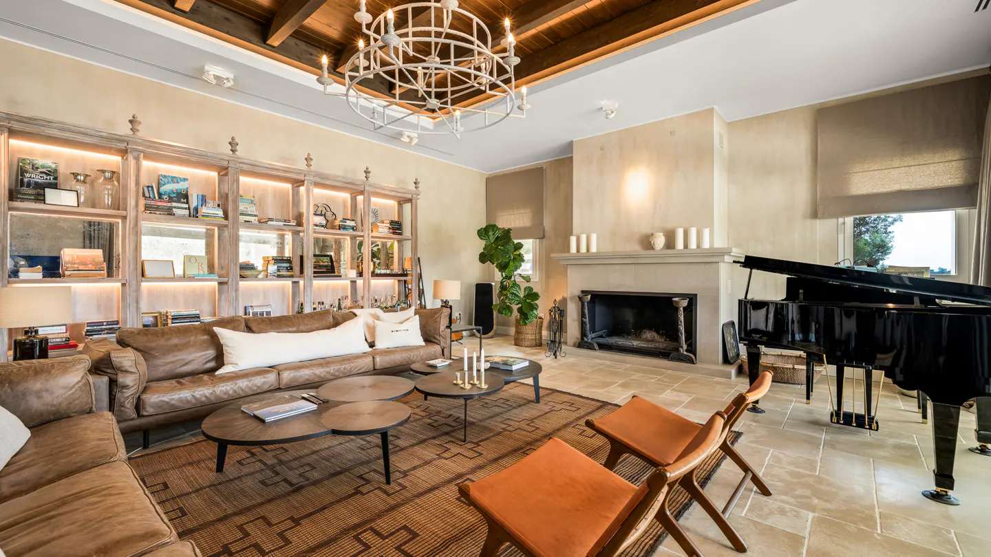 Living room with brown leather sofa, fireplace, piano, and bookshelf. A chandelier hangs from the wood-beamed ceiling.