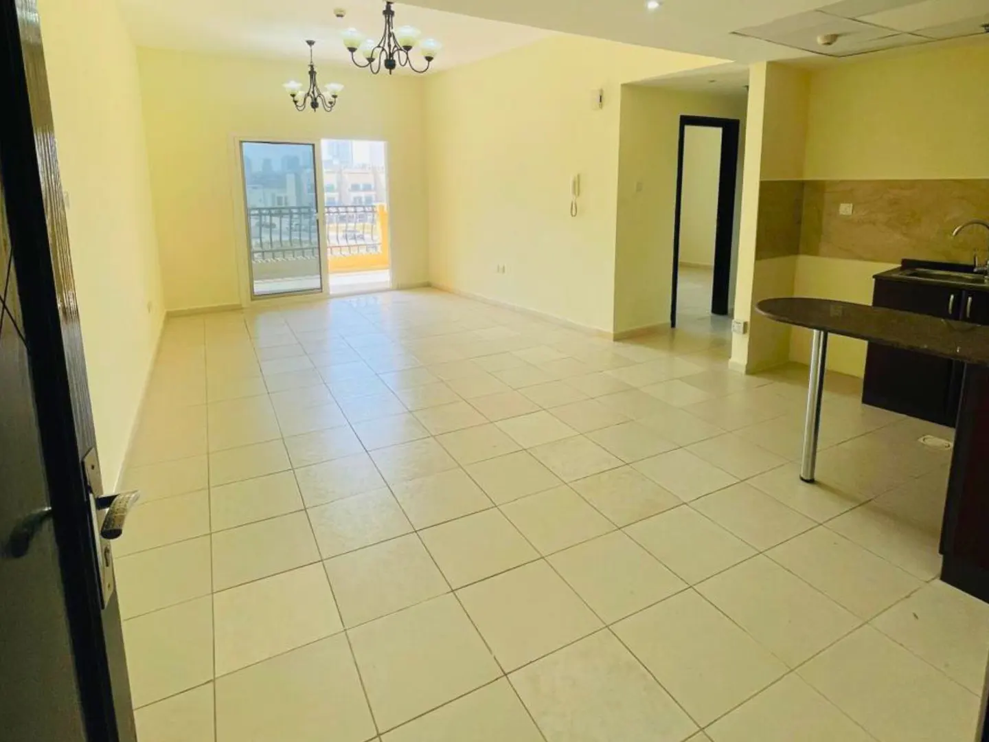 A bright, empty apartment with cream tile floors, yellow walls, and a balcony. A kitchen area is visible with dark cabinets and a breakfast bar.