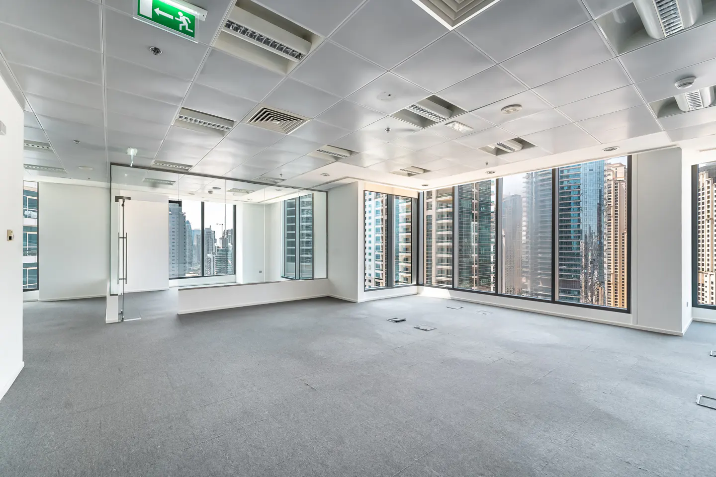 Empty office space with gray carpet, white walls, and large windows showing city buildings. An exit sign is visible on the ceiling.