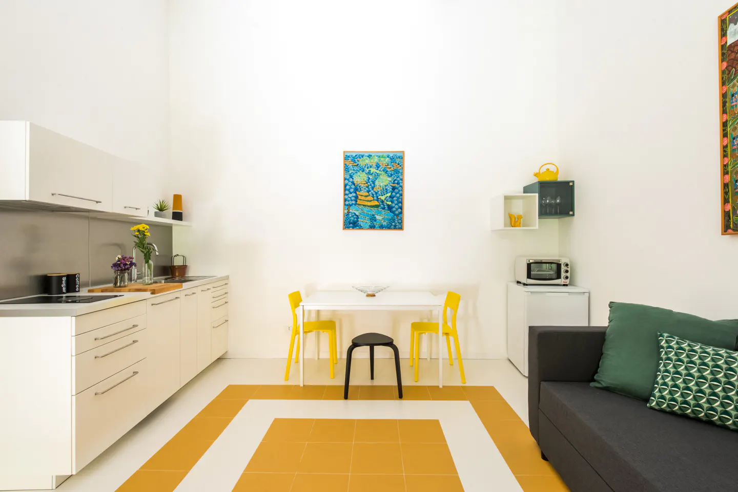 Bright, modern apartment interior with white walls, yellow accents, and a gray sofa. Kitchen cabinets line the left, dining table in the center.