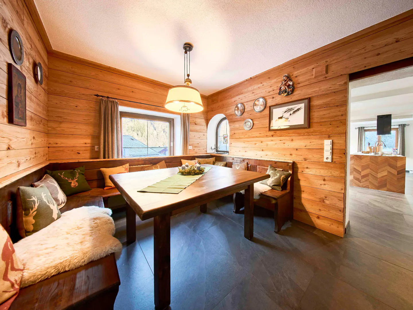 Cozy dining area with wood-paneled walls, a corner bench with pillows, and a wooden table with a green runner. A kitchen is visible through a doorway.