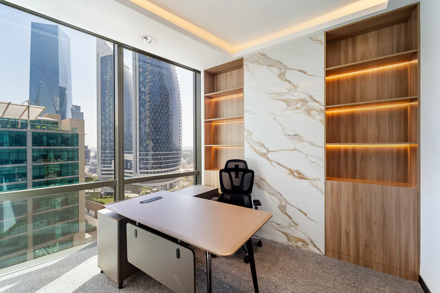 Bright office with a desk, chair, and built-in shelves. Large windows offer a city view with skyscrapers.