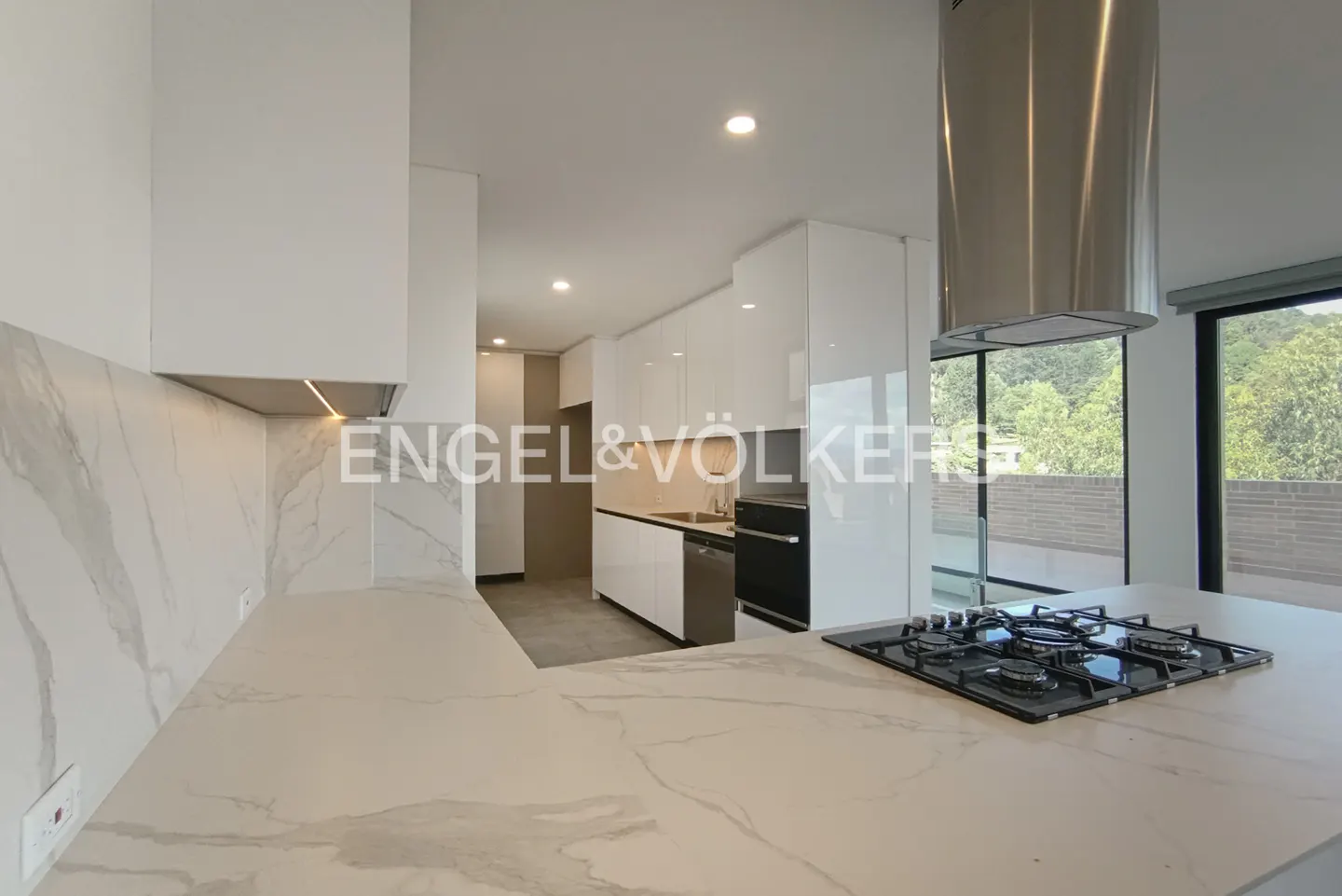 A modern kitchen with white cabinets, marble countertops, and stainless steel appliances. A gas stovetop sits on the island. Large windows offer a view of greenery.