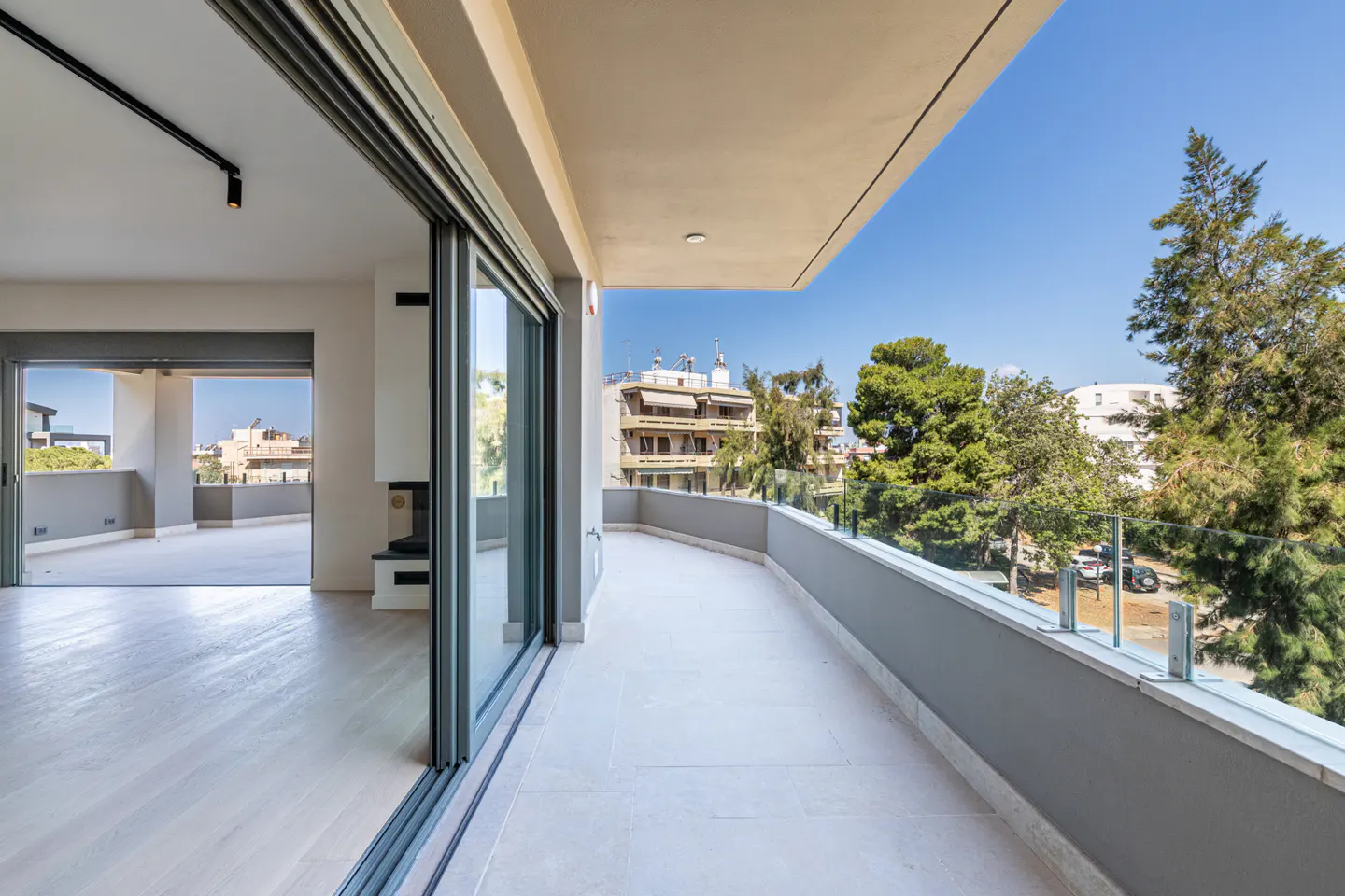 Spacious modern balcony with light stone flooring, glass railings, and a view of trees and buildings under a clear blue sky.