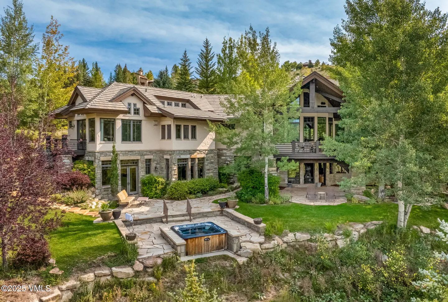 Exterior view of a large, multi-story home with a hot tub, stone patio, and lush green landscaping.
