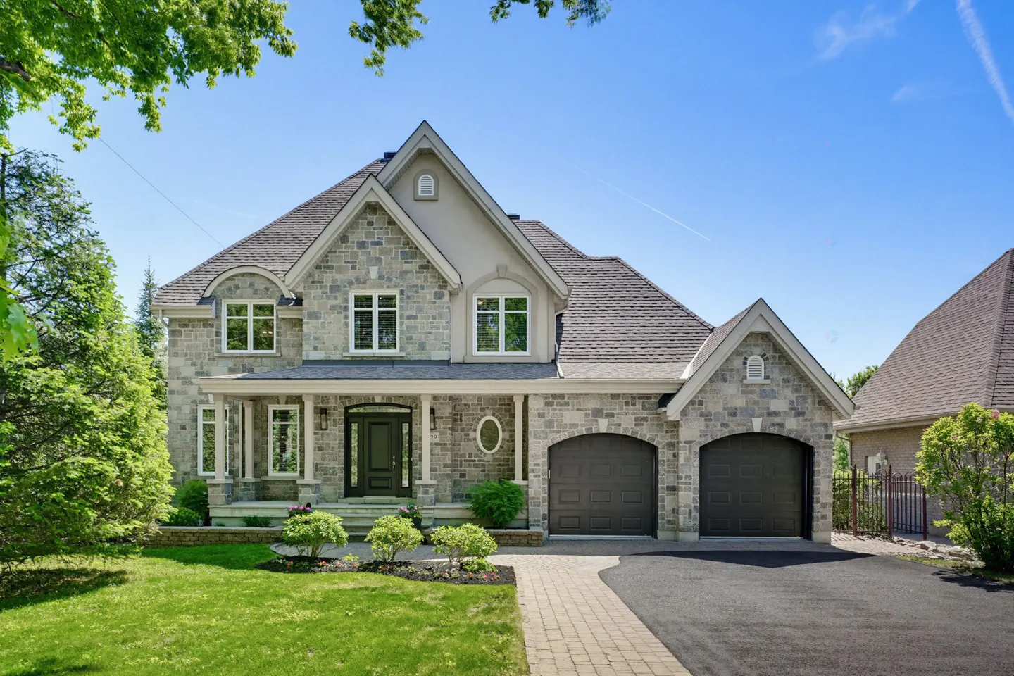 Two-story stone house with a dark roof, double garage, front porch, and manicured lawn under a clear blue sky.