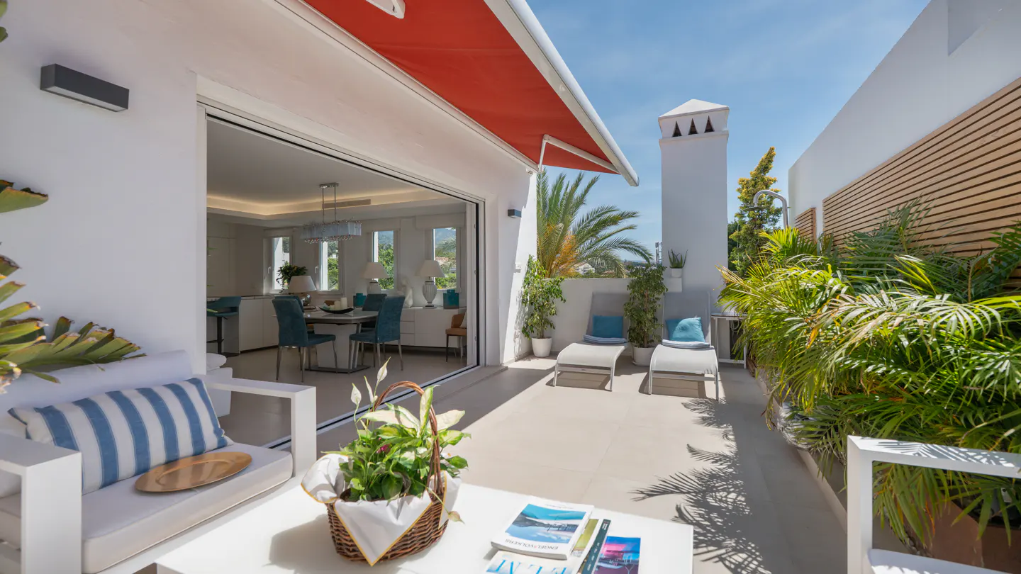Sunny terrace with white furniture, blue-striped cushions, two lounge chairs, lush plants, and a view into a dining room.