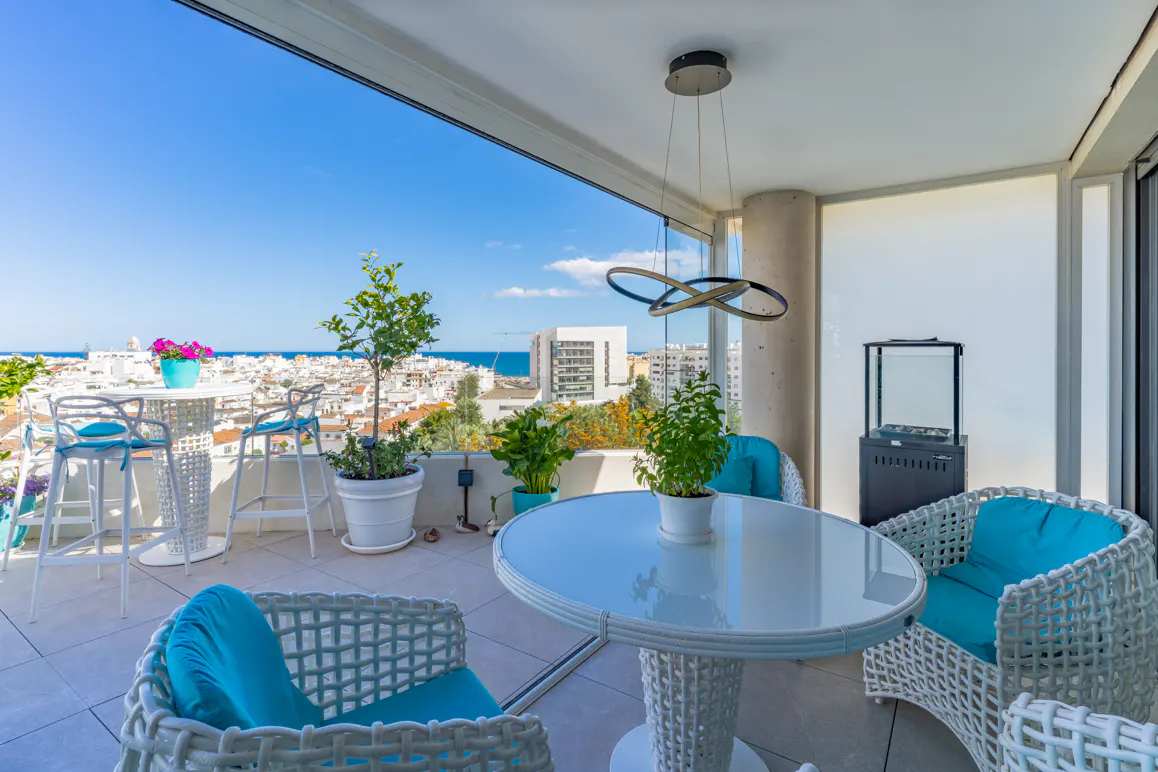 A bright, modern balcony with white wicker furniture, turquoise cushions, and potted plants overlooking a city and ocean view.