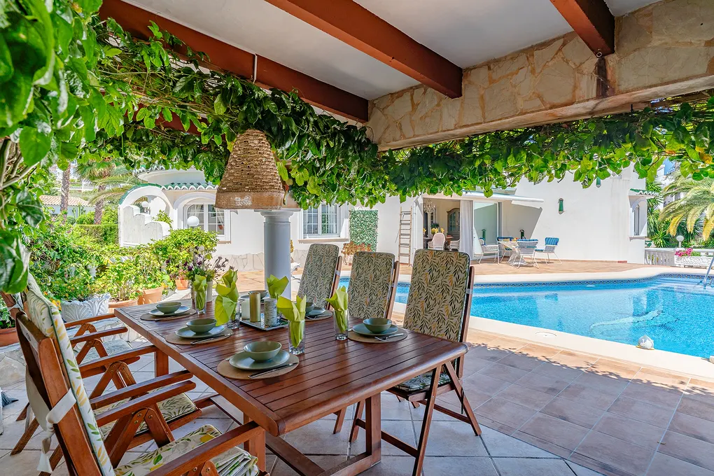 Outdoor dining area with a wooden table set for a meal, chairs, and a pool in the background. Green vines hang overhead.
