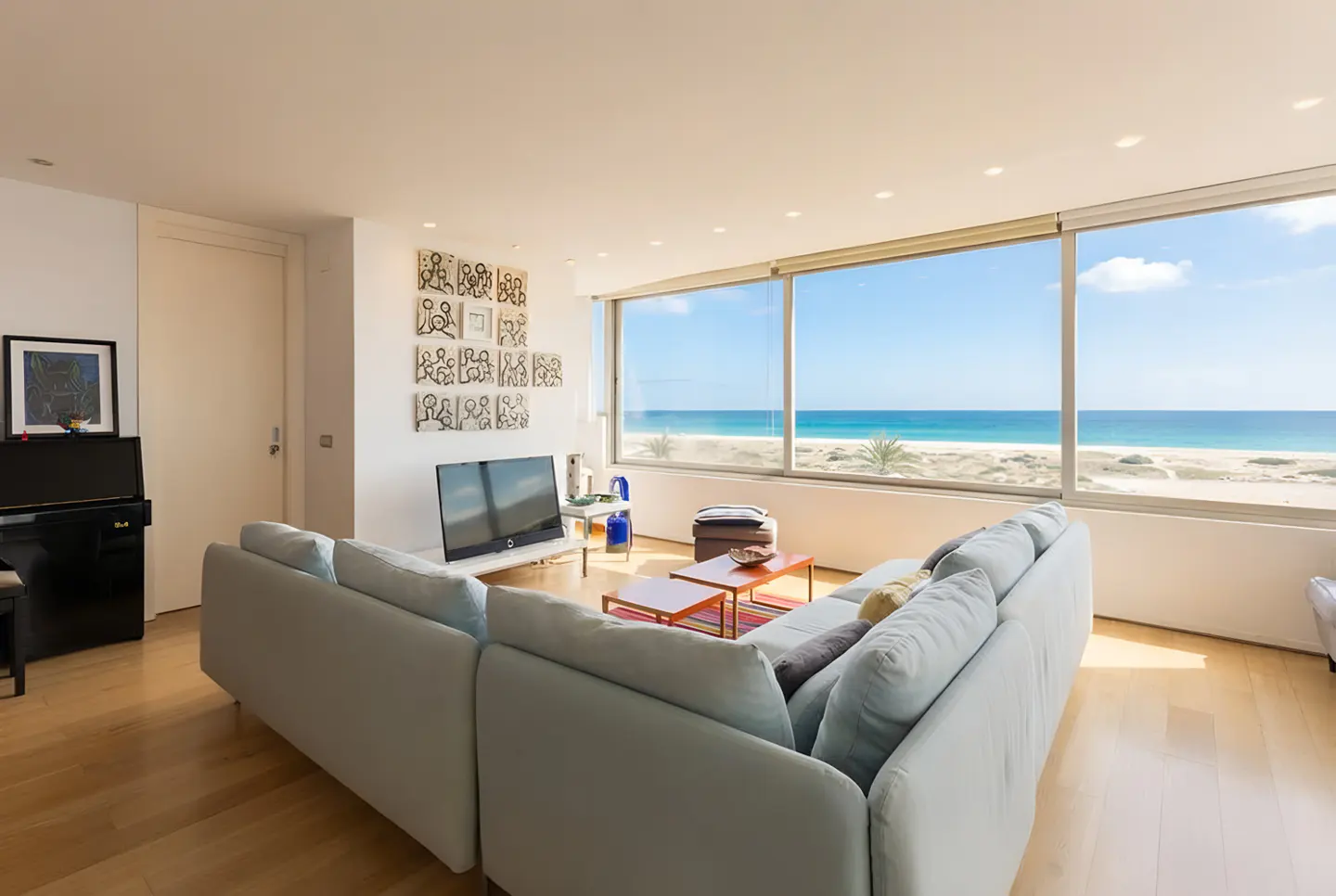 Bright living room with a large window view of the ocean. A light blue sofa faces a TV and artwork. A piano sits against the left wall.