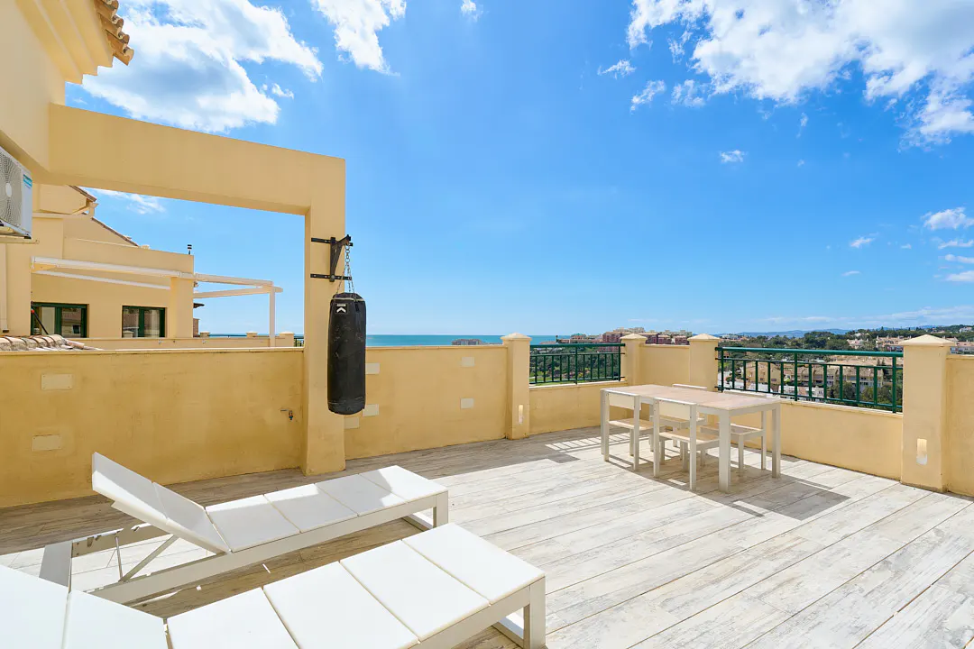 Rooftop patio with ocean view. Features a punching bag, table with chairs, and lounge chairs. Sunny blue sky.