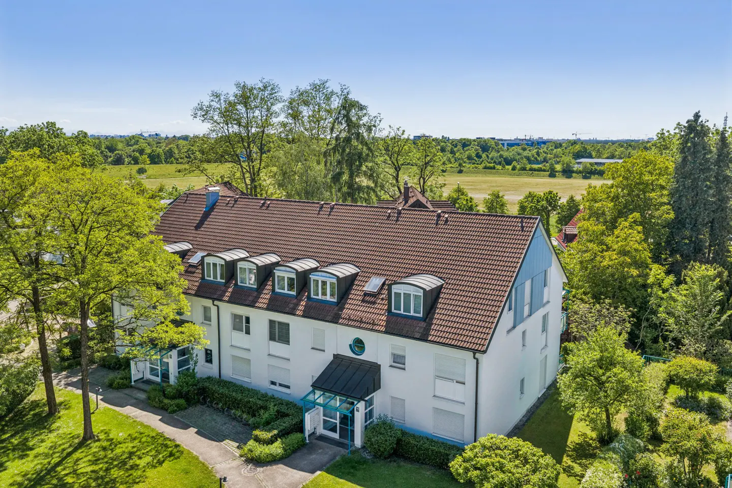Aerial view of a white multi-story apartment building with a brown tiled roof, surrounded by lush green trees and grass.