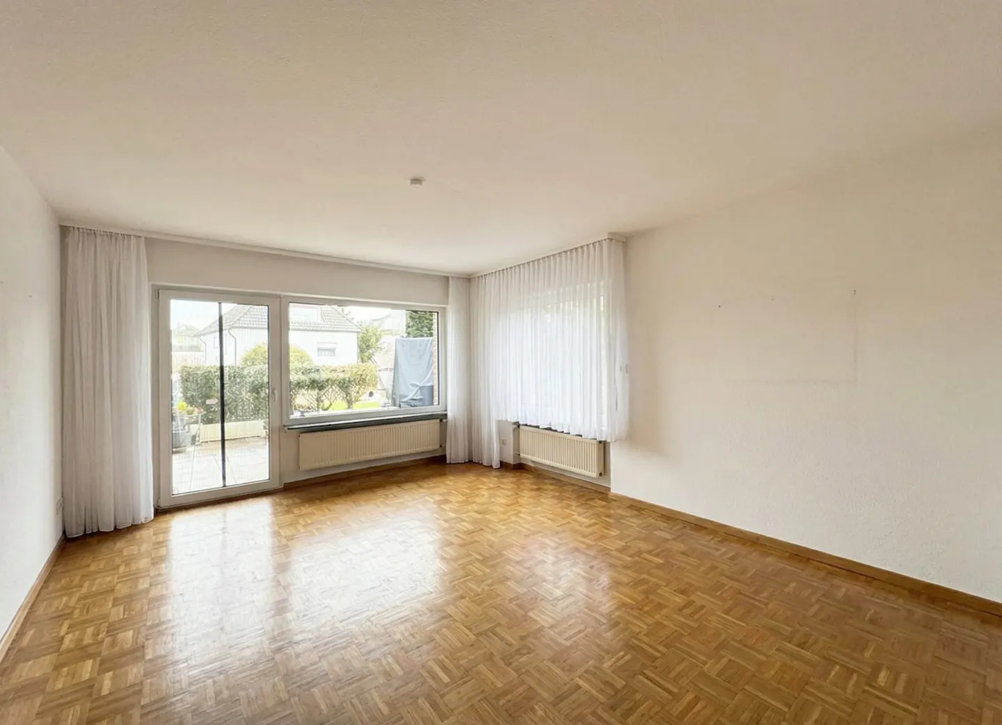 Empty room with parquet floor, white walls, and large windows with white curtains. A radiator is under each window. Outside is a garden.