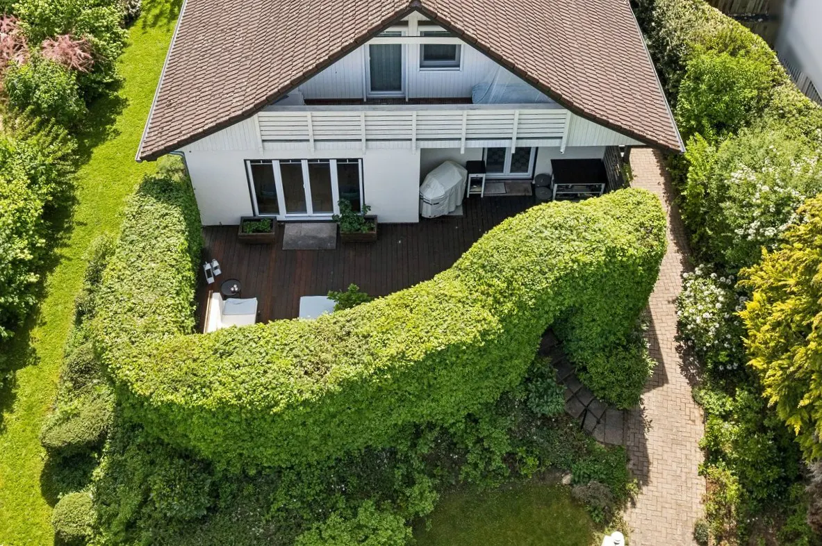 Aerial view of a white house with a brown roof, surrounded by a tall, green hedge and lush greenery. A wooden deck and patio furniture are visible.