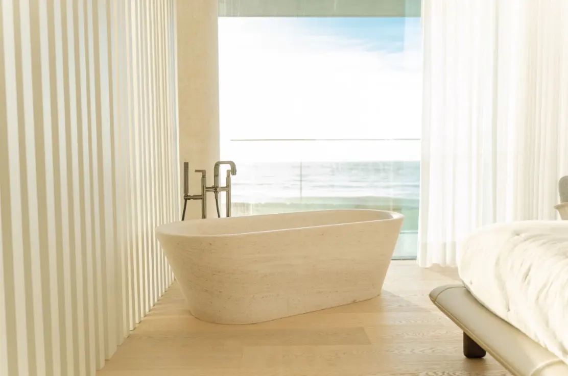 Modern bathroom with a stone tub and chrome faucet, next to a large window with ocean view. Neutral color palette.