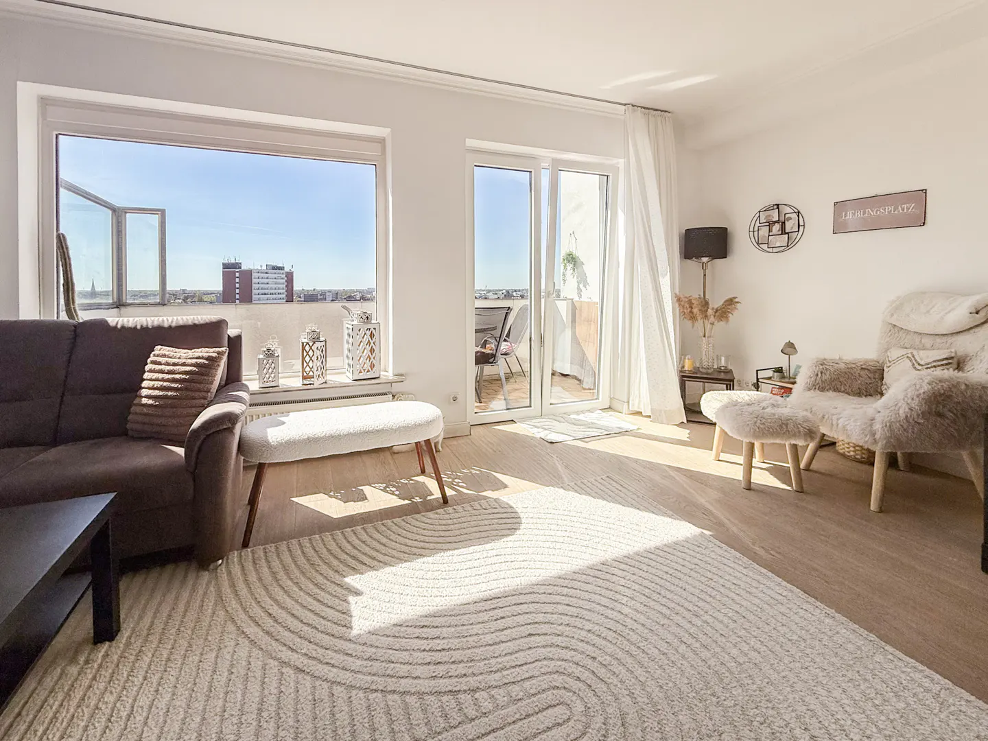 Bright living room with brown sofa, white rug, and balcony access. Sunlight streams through the window, illuminating the wooden floor.
