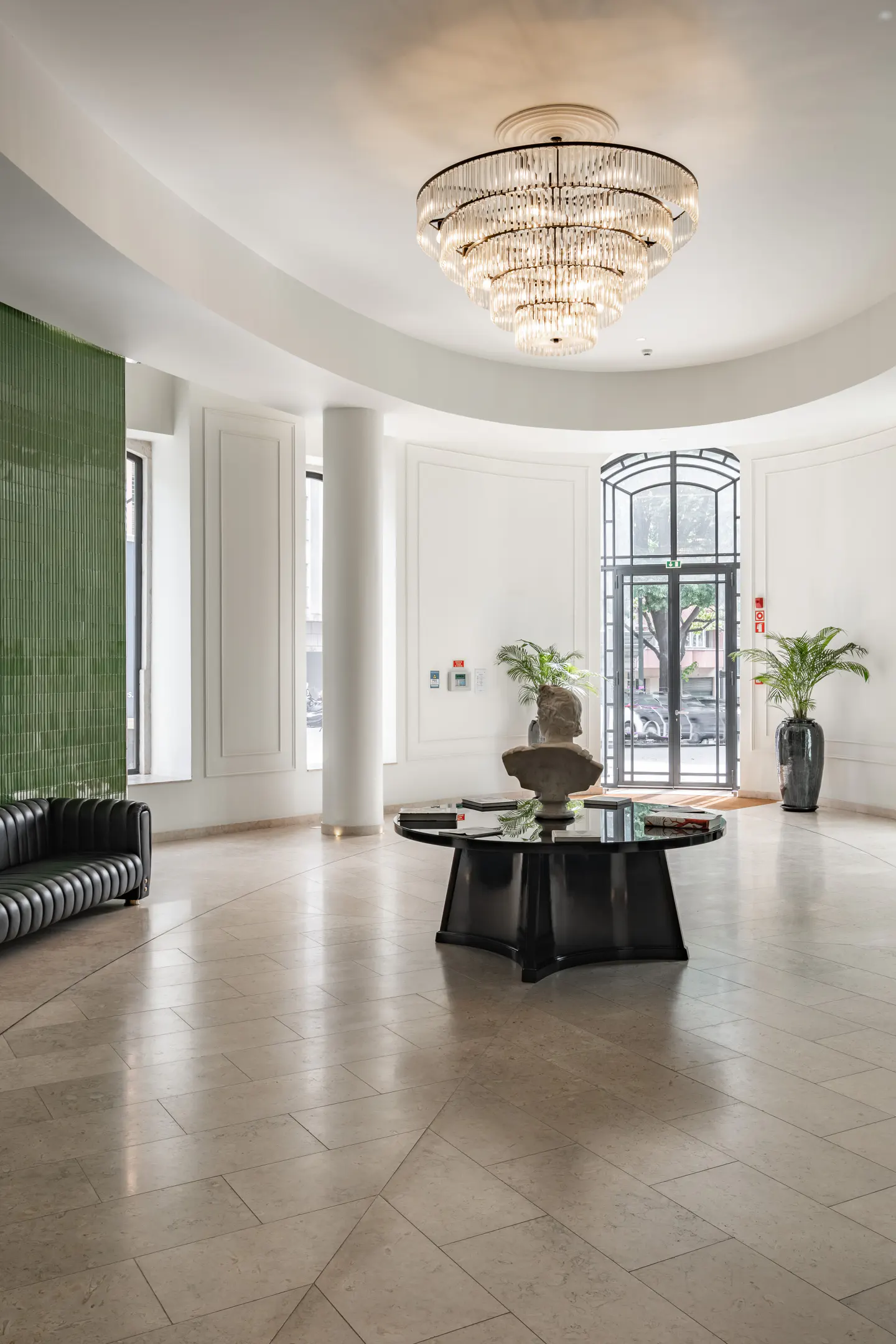 Bright lobby with a crystal chandelier, black table with plants, and a black leather sofa. The floor is light beige tile.