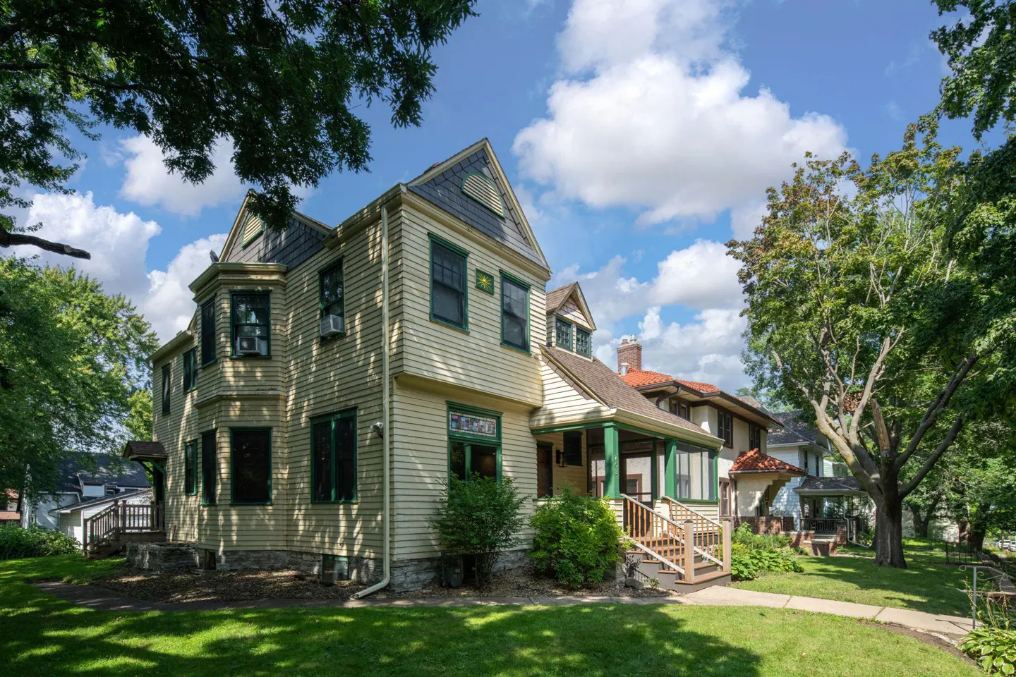 A two-story, light yellow house with green trim and a gray roof on a green lawn under a blue sky with white clouds.