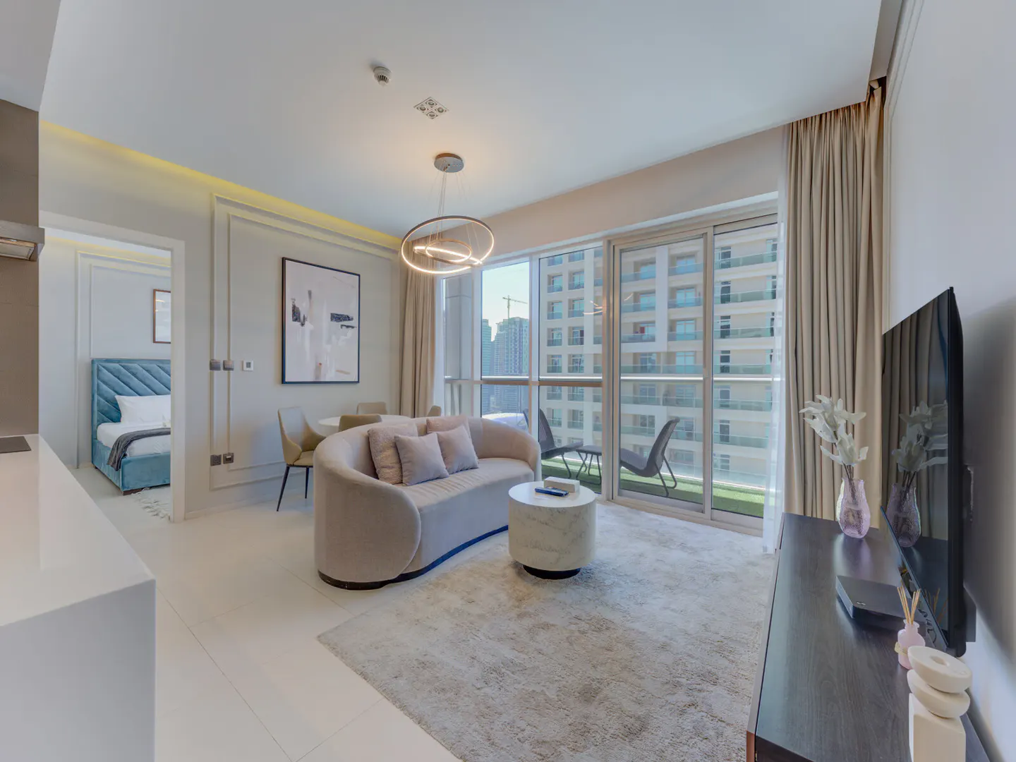 Bright apartment interior with a curved sofa, round table, and balcony view of city buildings. A bedroom is visible through a doorway.