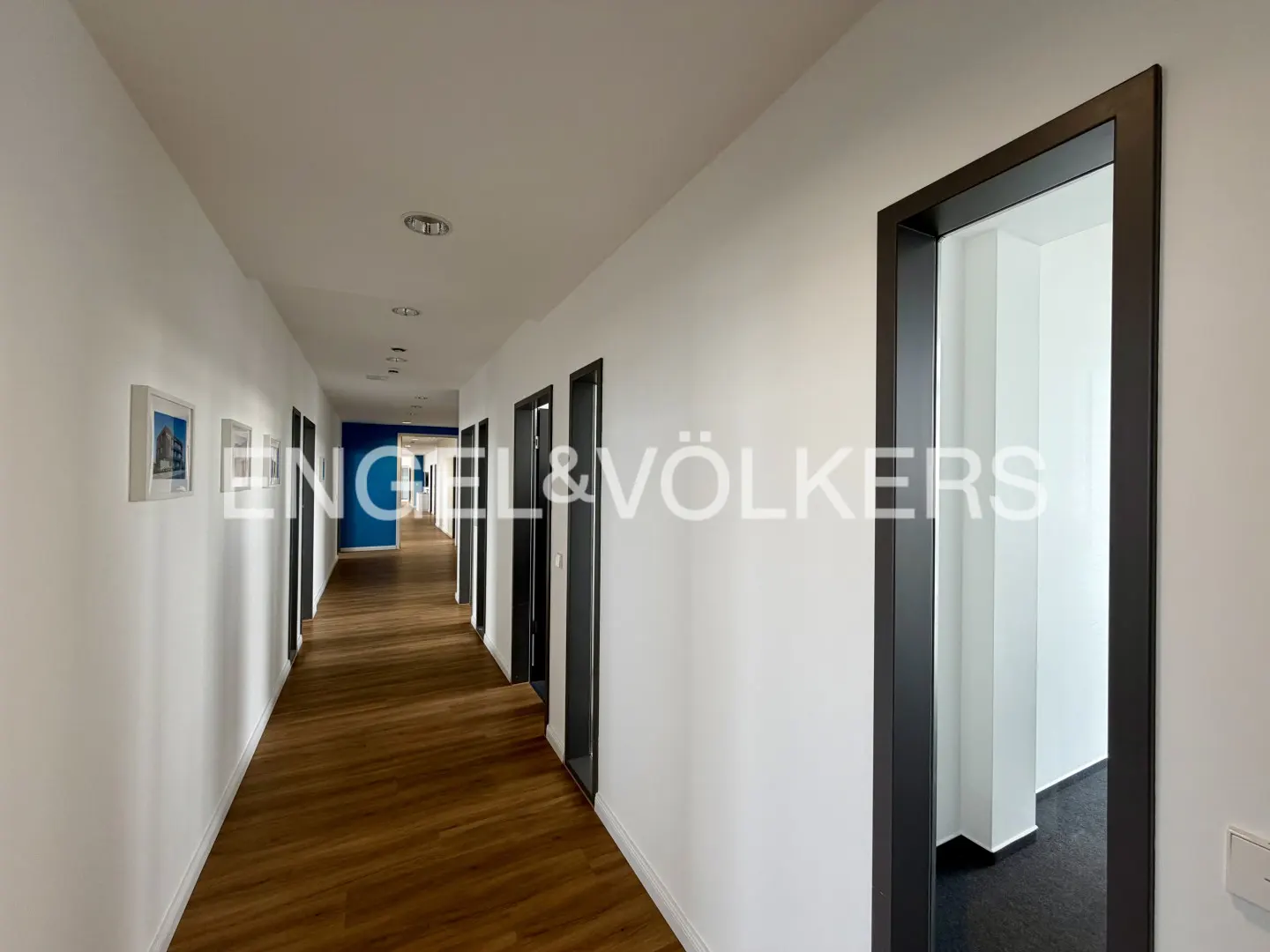Long hallway with wood floors, white walls, and black door frames. Engel & Völkers logo on the wall. Blue accent wall at the end.