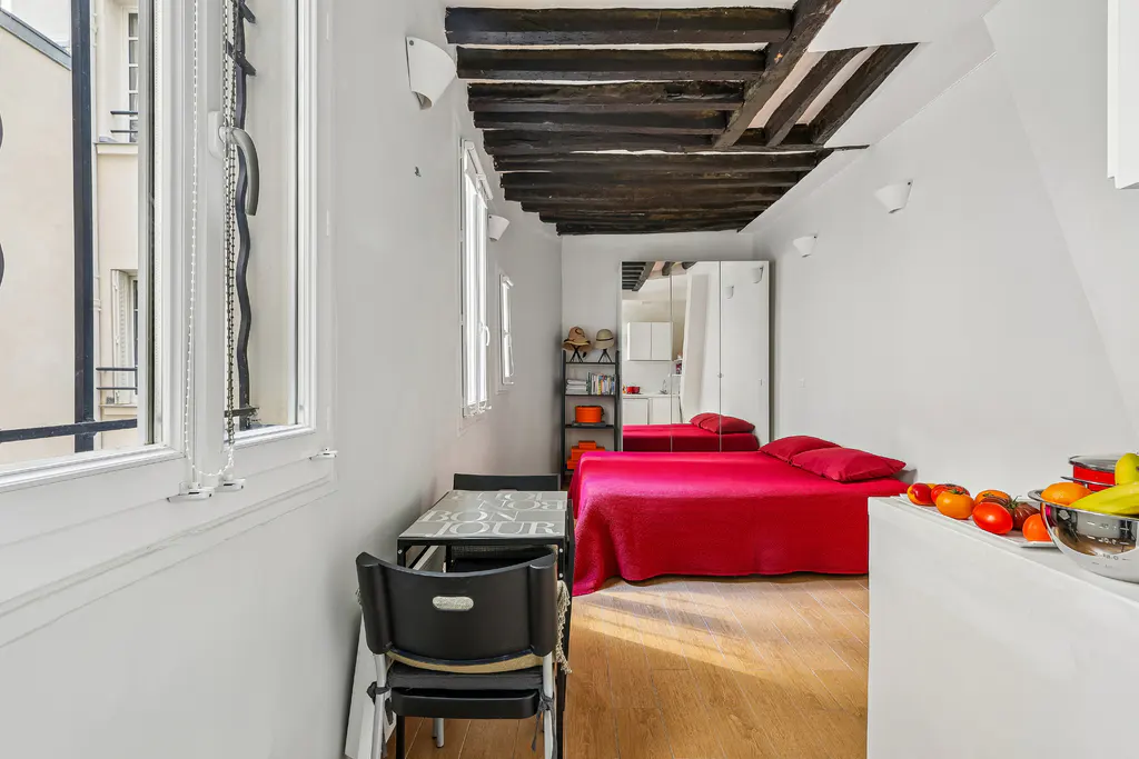 A compact studio apartment with a red bed, wooden floors, and exposed dark wood beams on the ceiling. A table and chairs are near a window.