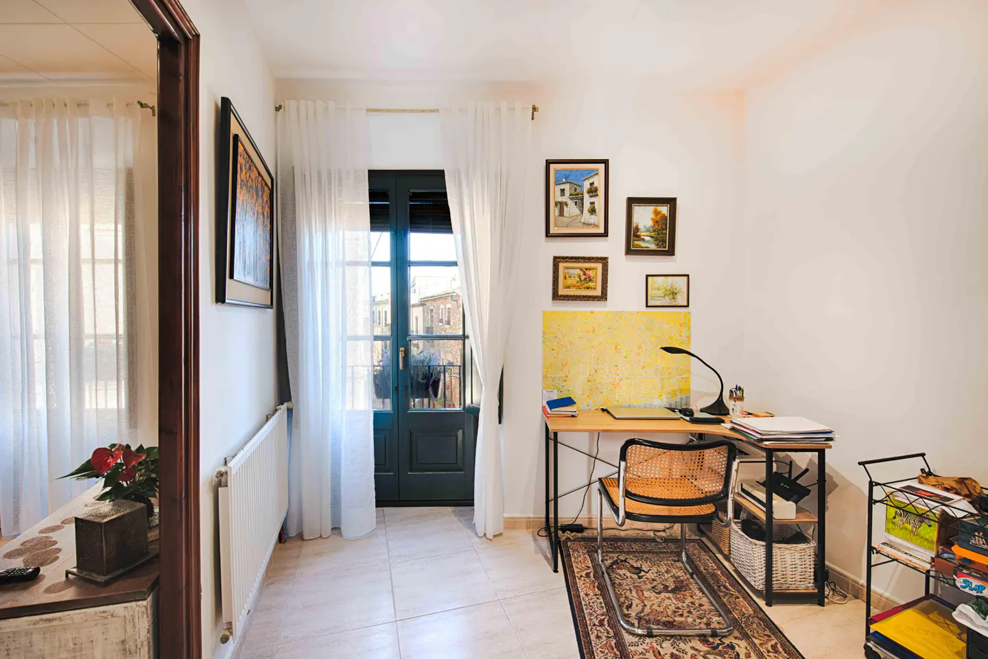 A bright room with a desk, chair, and balcony door. Paintings hang on the white wall above a yellow map. A rug sits under the chair.