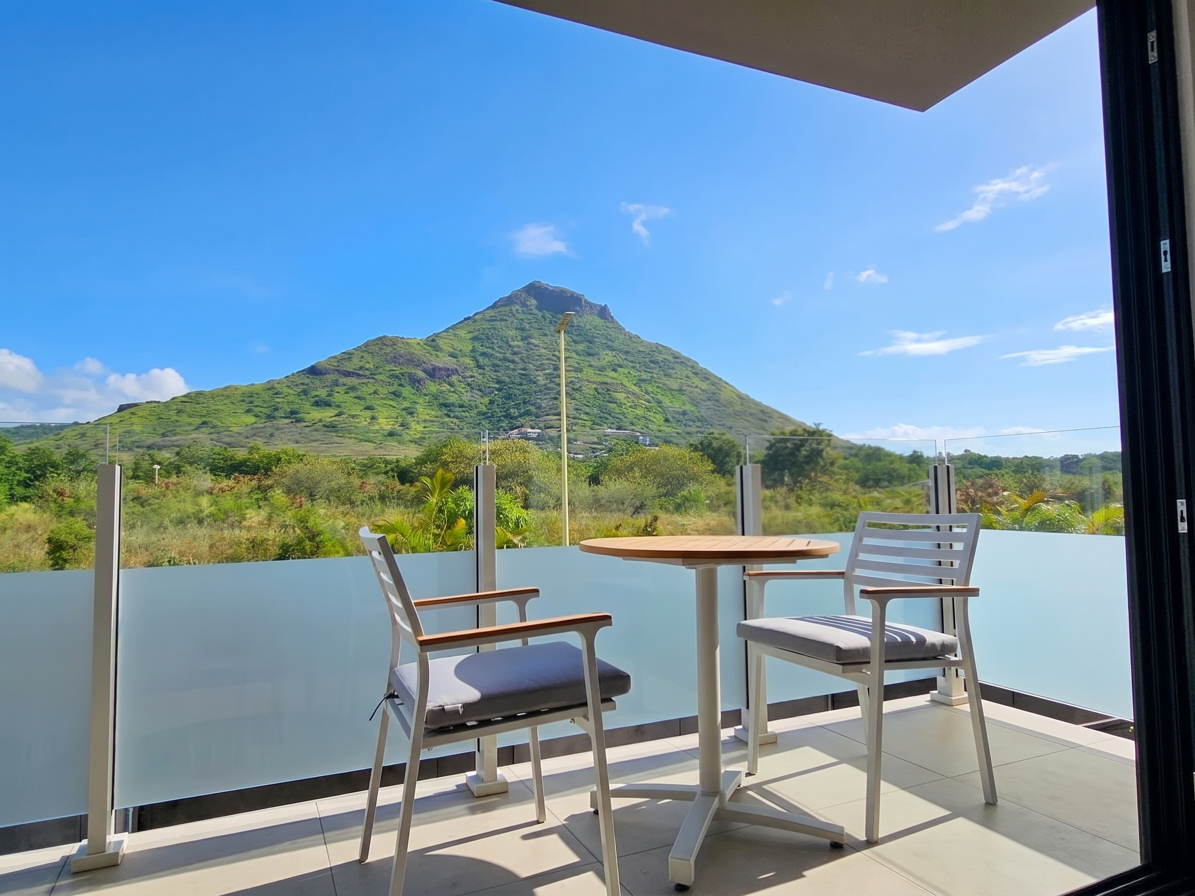 Balcony view with table and chairs overlooking a green mountain under a blue sky.