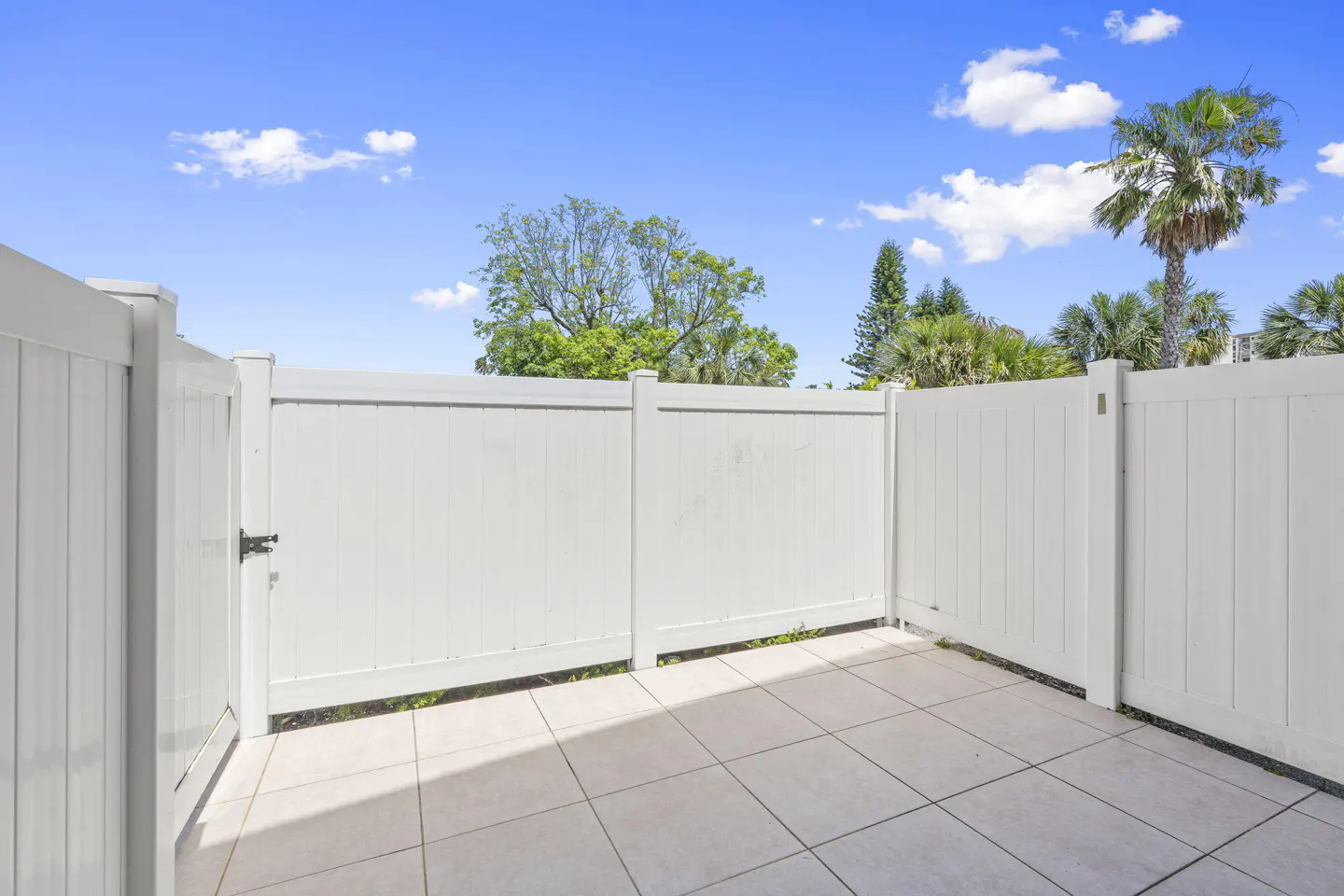 A fenced patio with white tile flooring, surrounded by a white vinyl fence. Trees and a blue sky with clouds are visible above the fence.