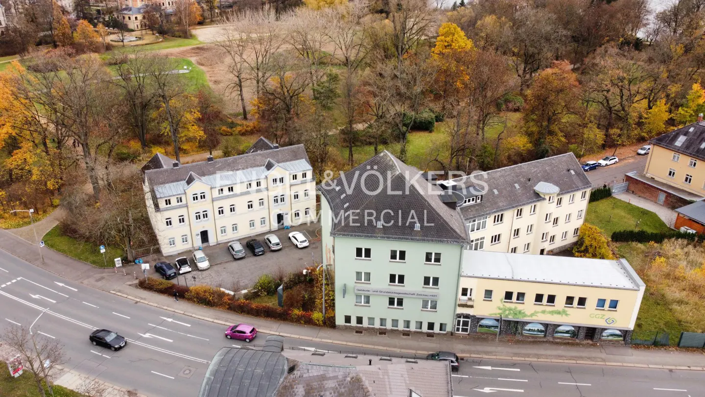Aerial view of a multi-building commercial property with parking, trees, and a road with cars. Engel & Völkers logo visible.