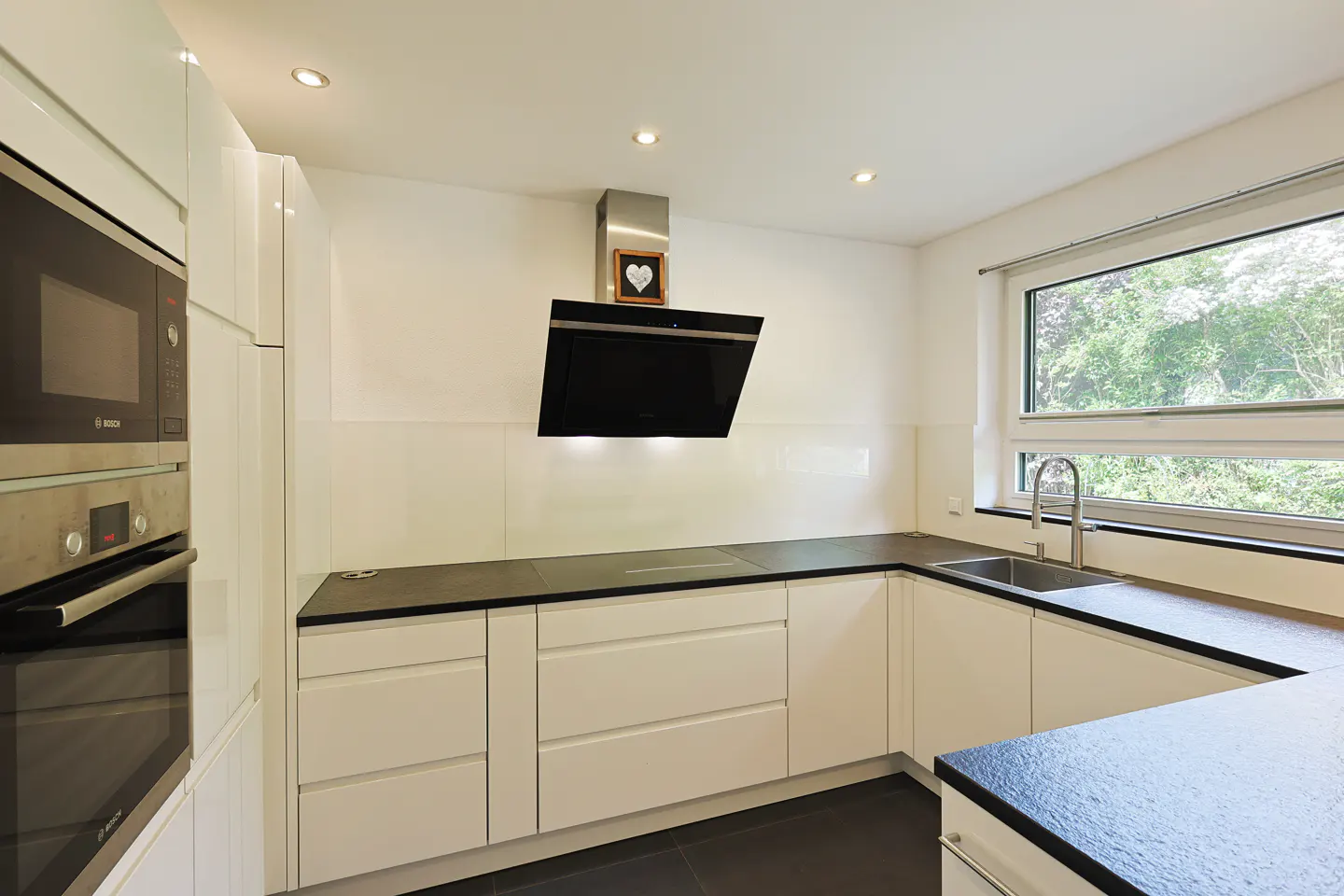 Modern kitchen with white cabinets, black countertops, built-in appliances, and a window overlooking greenery.