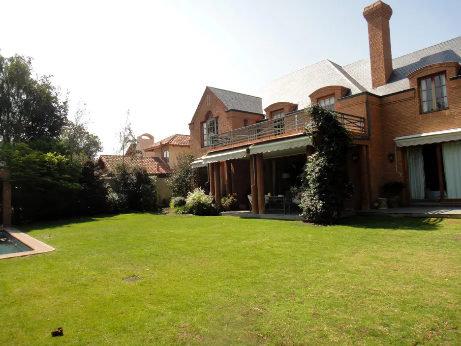 Exterior view of a large brick house with a green lawn, trees, and a pool in the background.