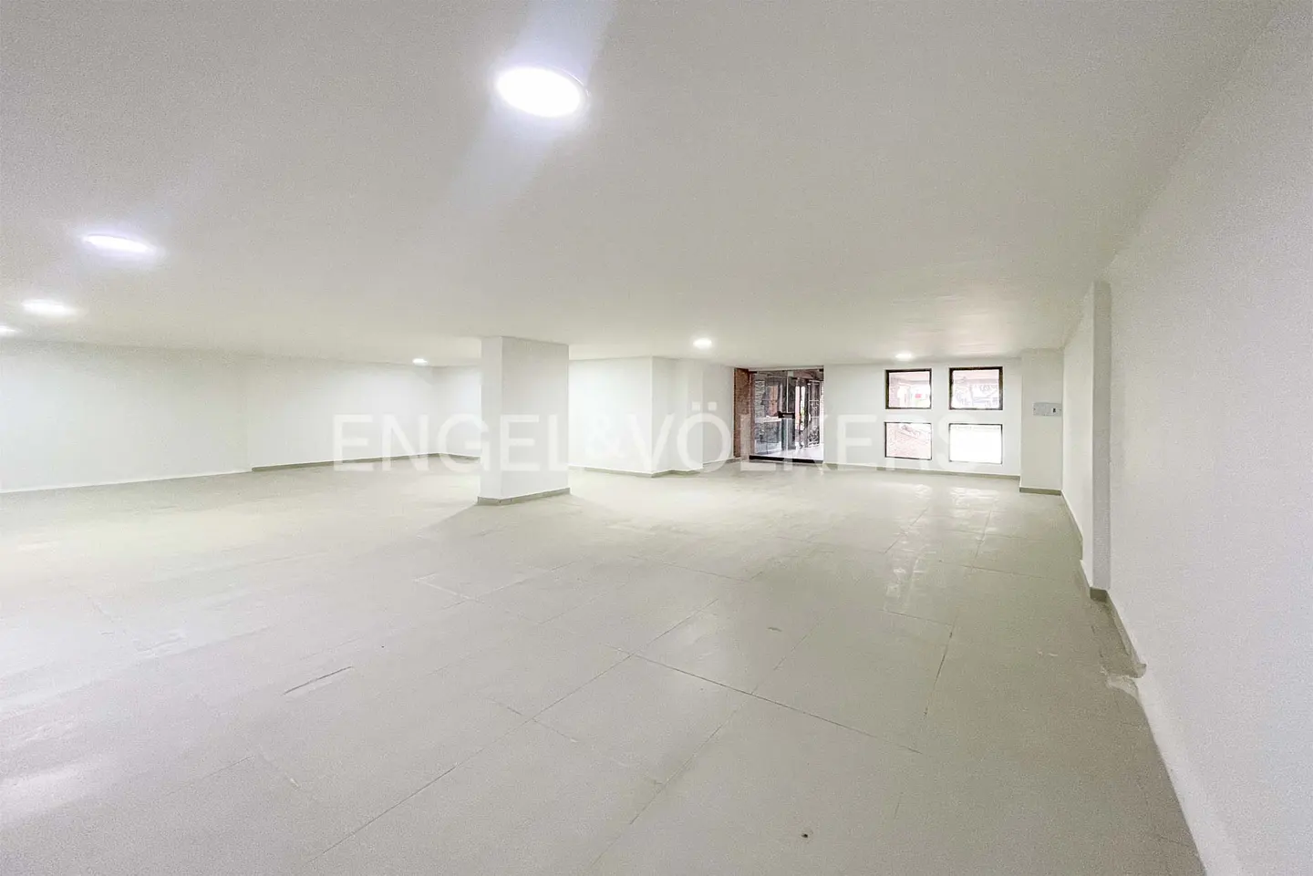 Bright, empty room with white walls, gray tile floor, and recessed lighting. A glass door and windows are visible in the background.