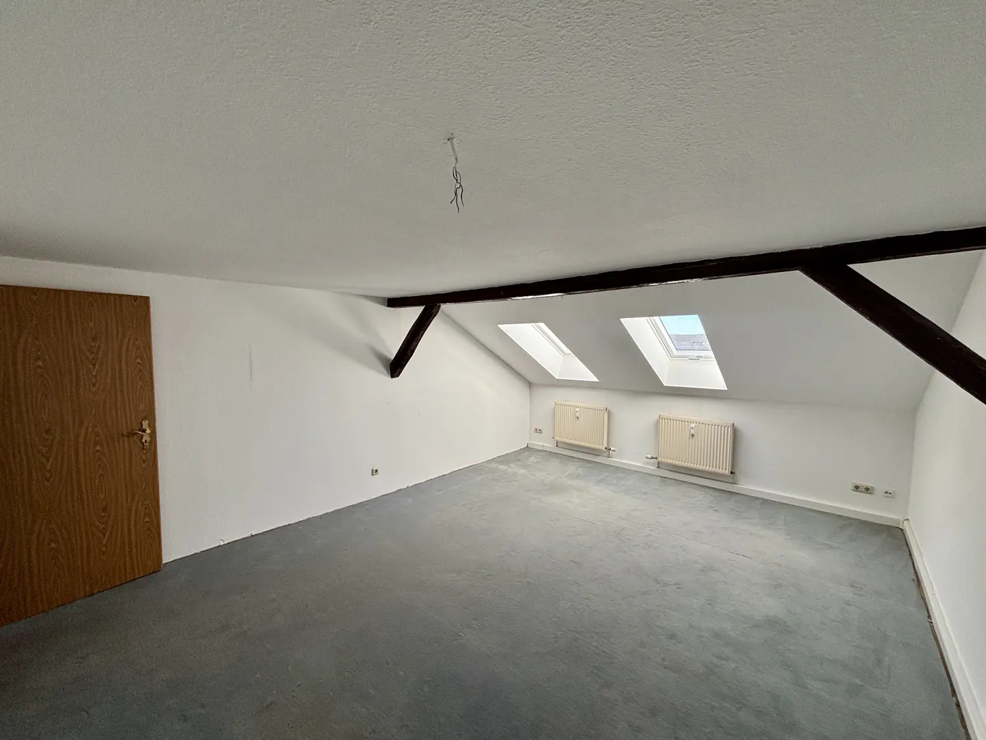 Attic room with gray carpet, white walls, and two skylights. A wooden door is on the left, and dark beams cross the ceiling.
