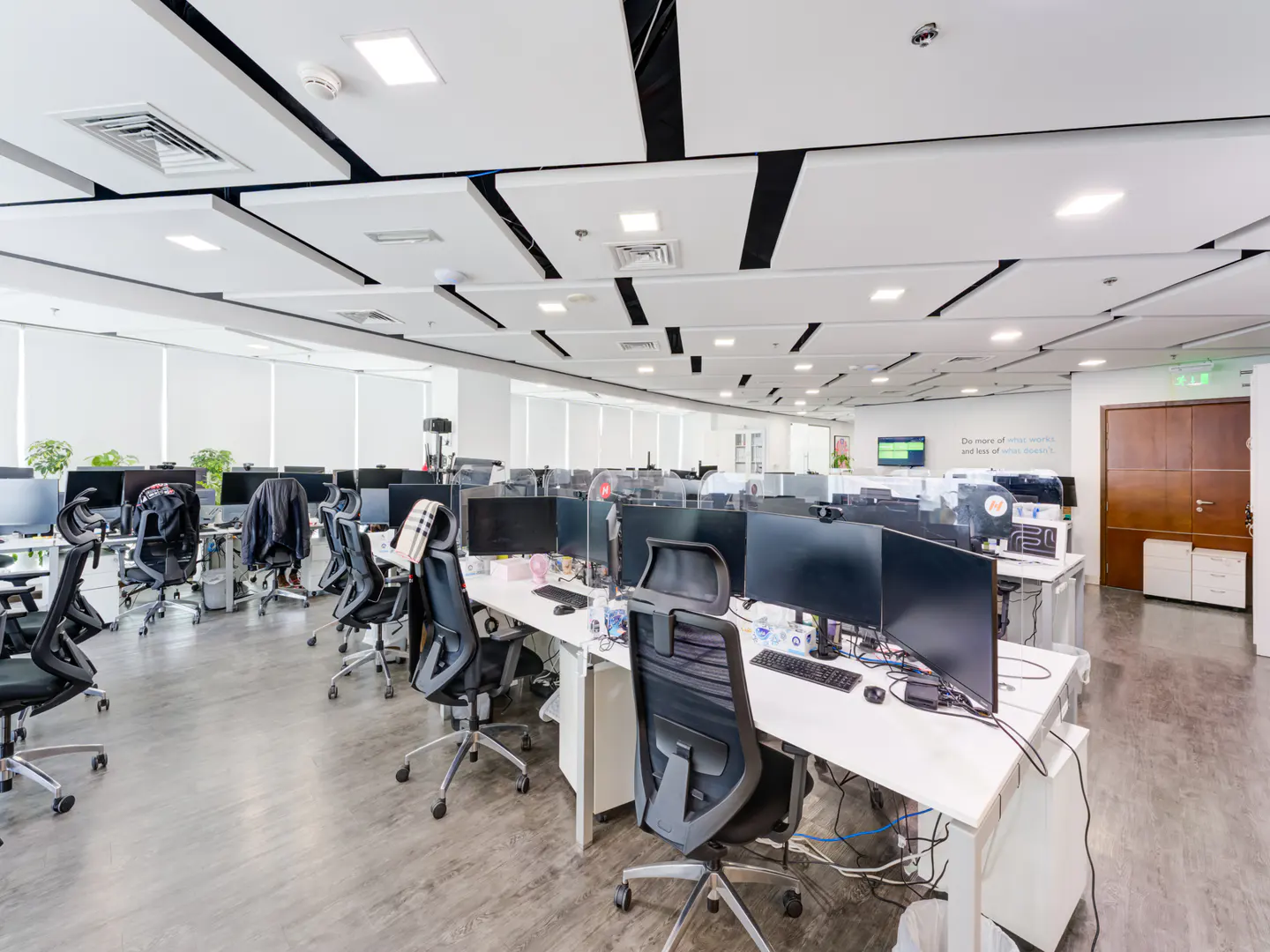 Bright, modern office space with rows of desks, black chairs, and computer monitors. White ceiling panels with black accents. Gray flooring.