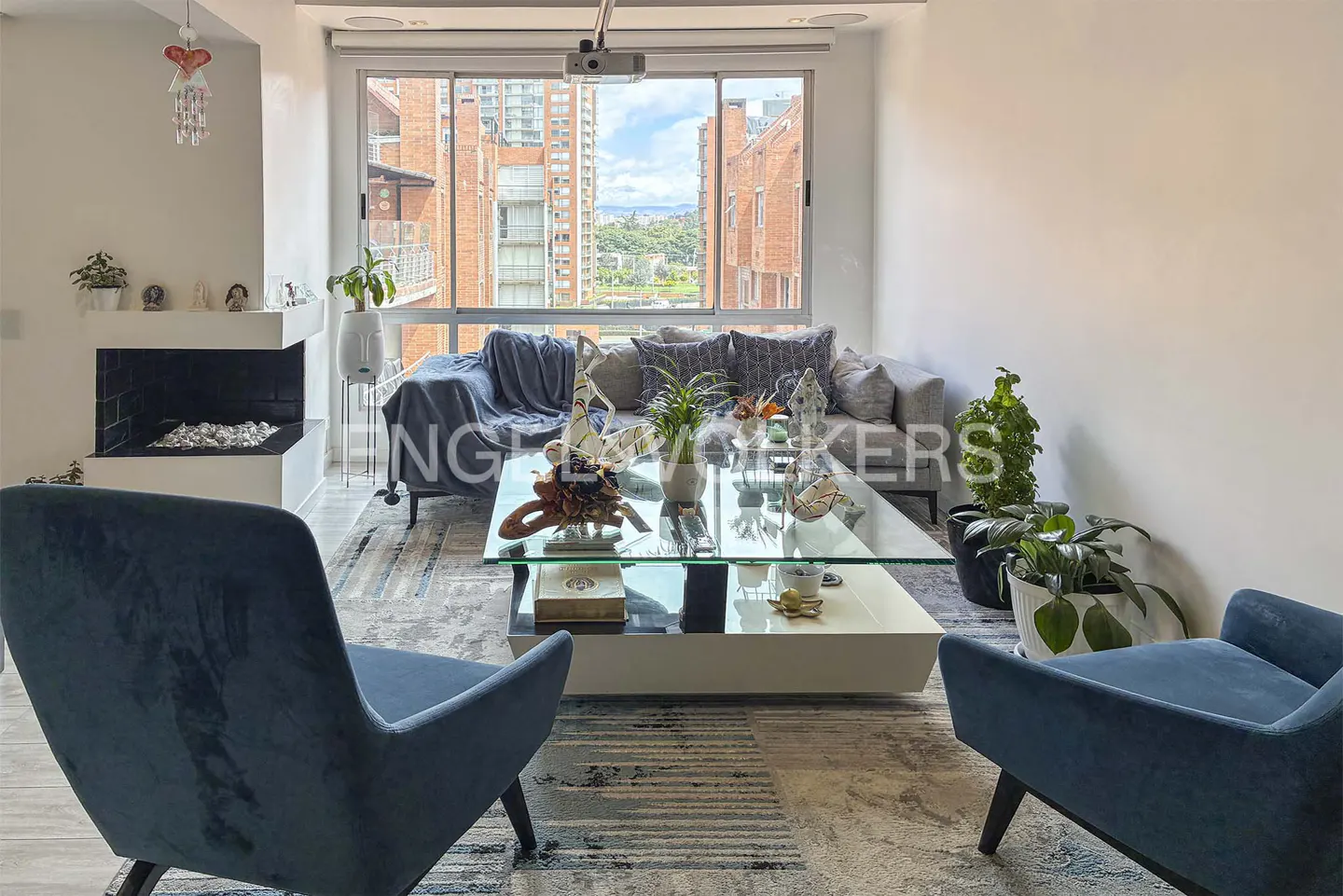 Modern living room with two blue armchairs, a glass coffee table, a grey sofa, and large windows overlooking city buildings.