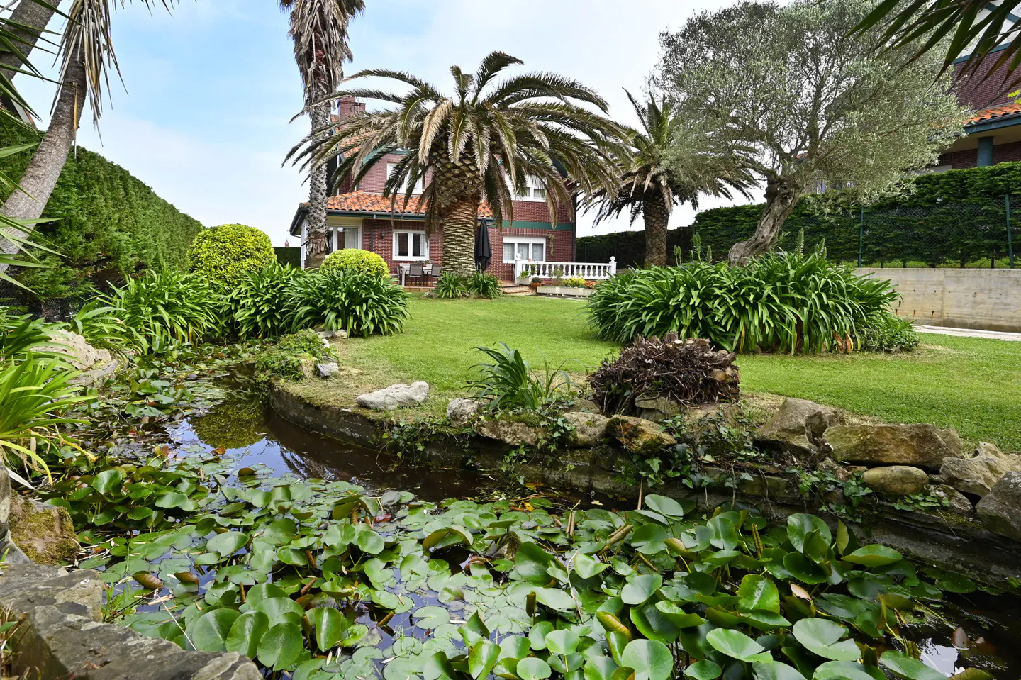 A lush garden with a pond full of lily pads in front of a brick house with palm trees on a sunny day.