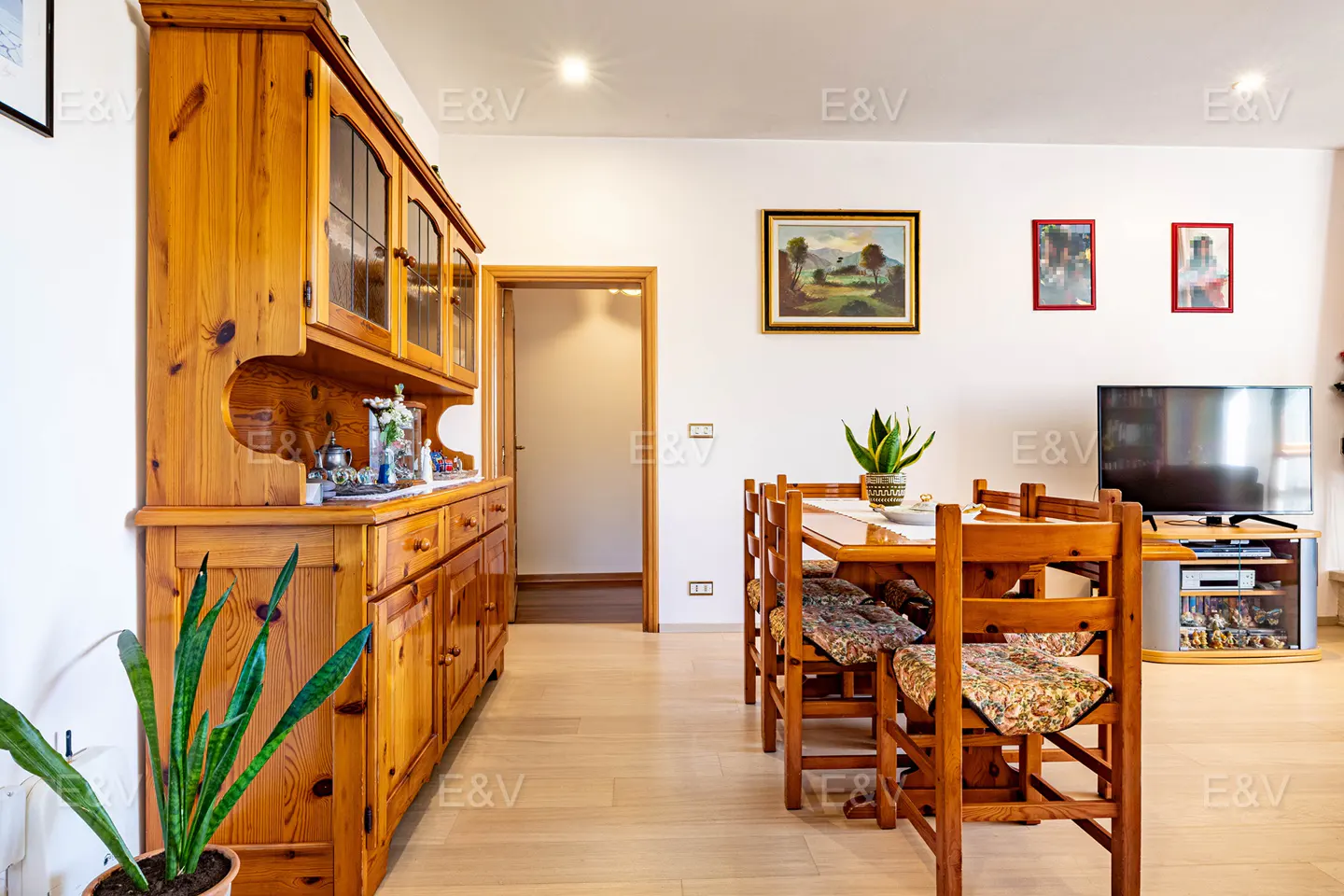 A dining room with a wooden table, chairs with floral cushions, and a large wooden hutch. Paintings adorn the white walls.