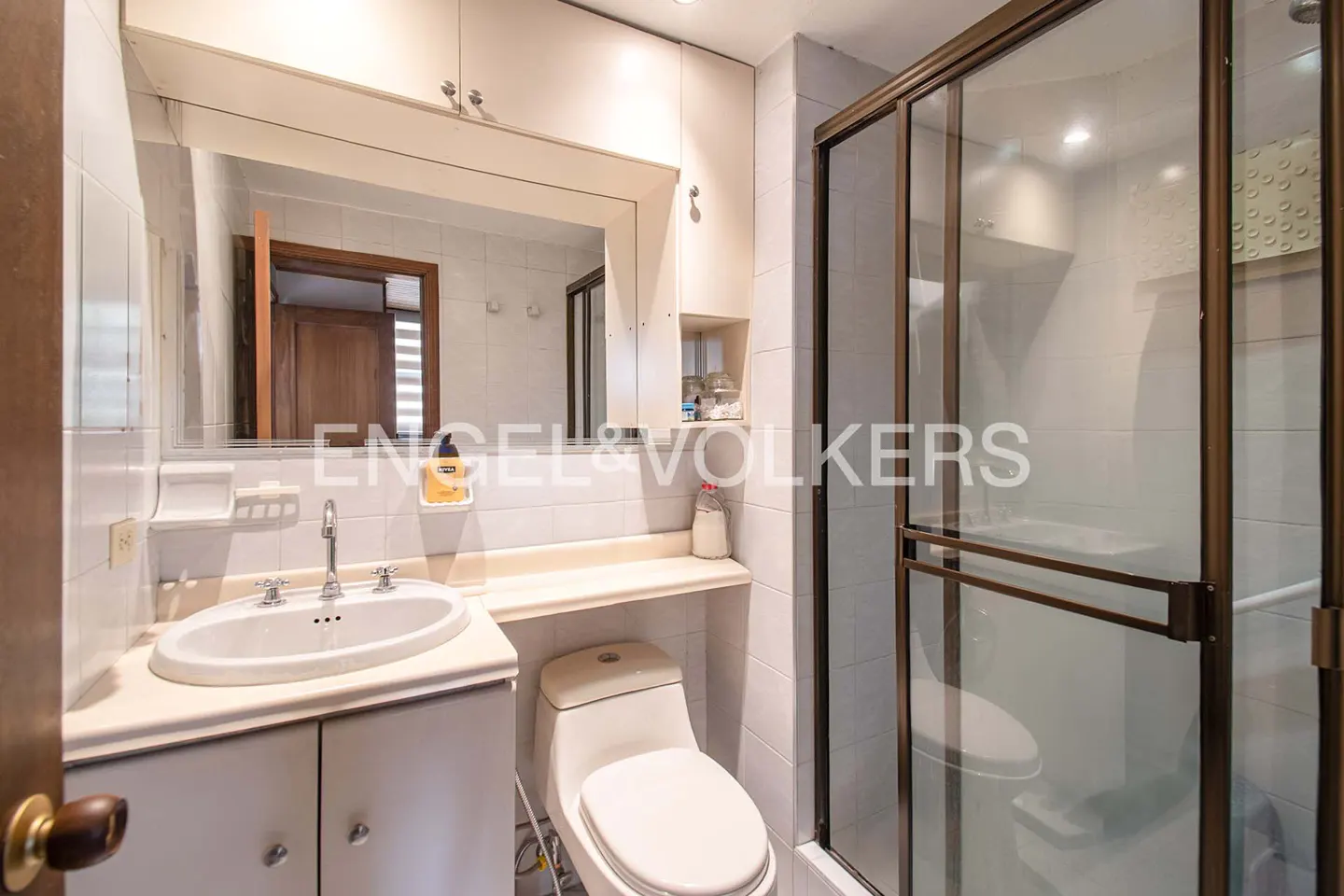 Bright bathroom with white tile, a sink with a mirror, a toilet, and a glass shower with bronze trim.