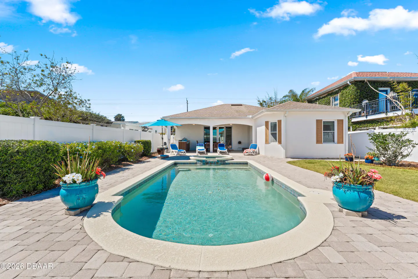 Backyard view of a pool with lounge chairs, a blue umbrella, and a white house on a sunny day.