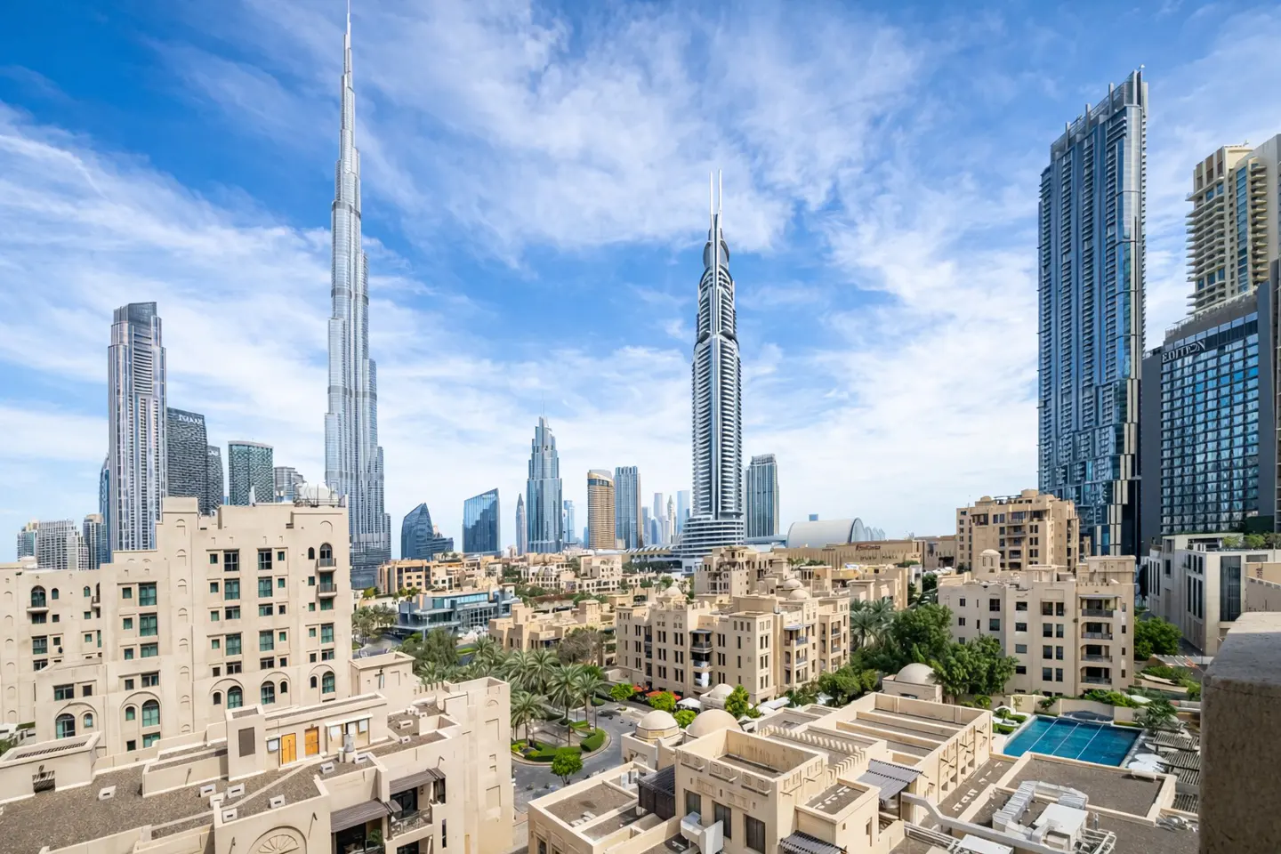 Dubai skyline view with Burj Khalifa, modern buildings, and beige residential complexes under a blue sky with clouds.