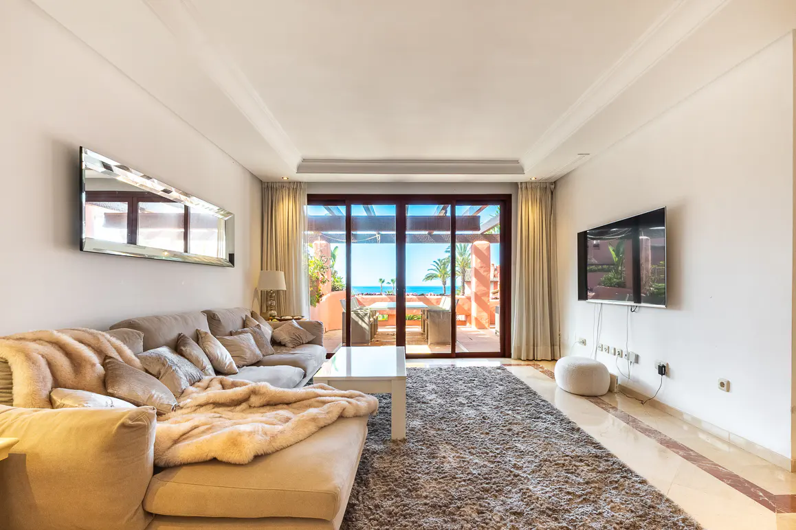 Living room with beige sofa, white walls, and a large window showing a balcony with ocean view. A TV hangs on the wall.