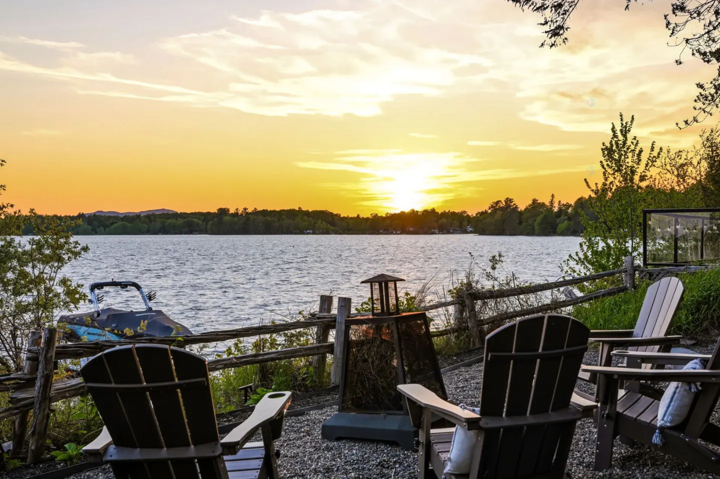 Lake view with Adirondack chairs and fire pit at sunset. Boat parked behind a wooden fence.