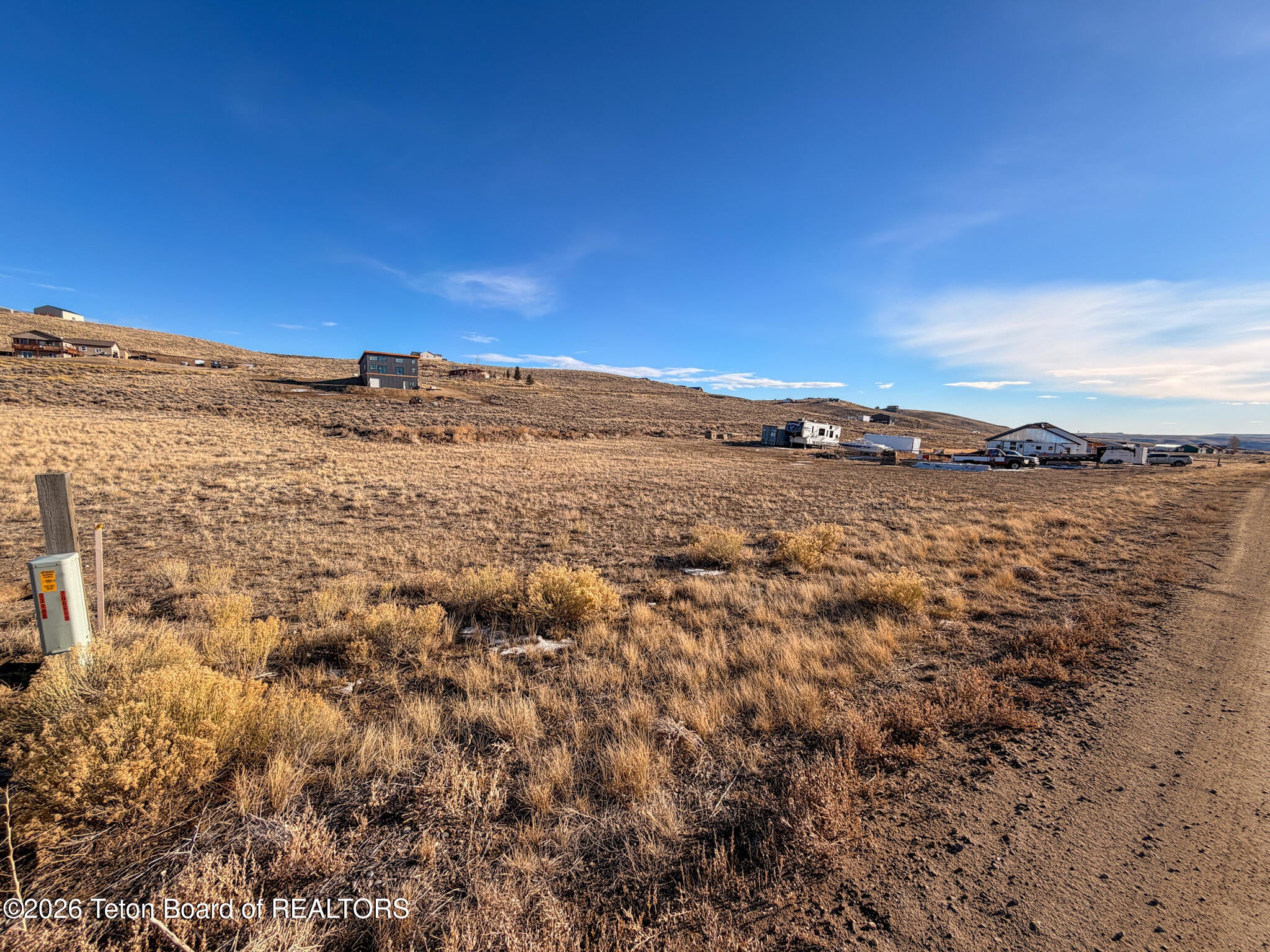 A dirt road borders a vast, dry, grassy lot under a clear blue sky, with distant houses on a rolling hillside. [AI-generated text]