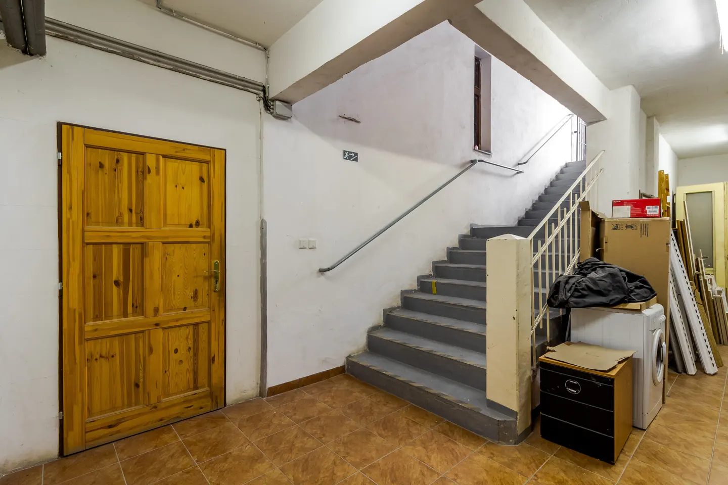 A wooden door next to a grey staircase in a hallway with tiled floors, storage boxes, and a washing machine.