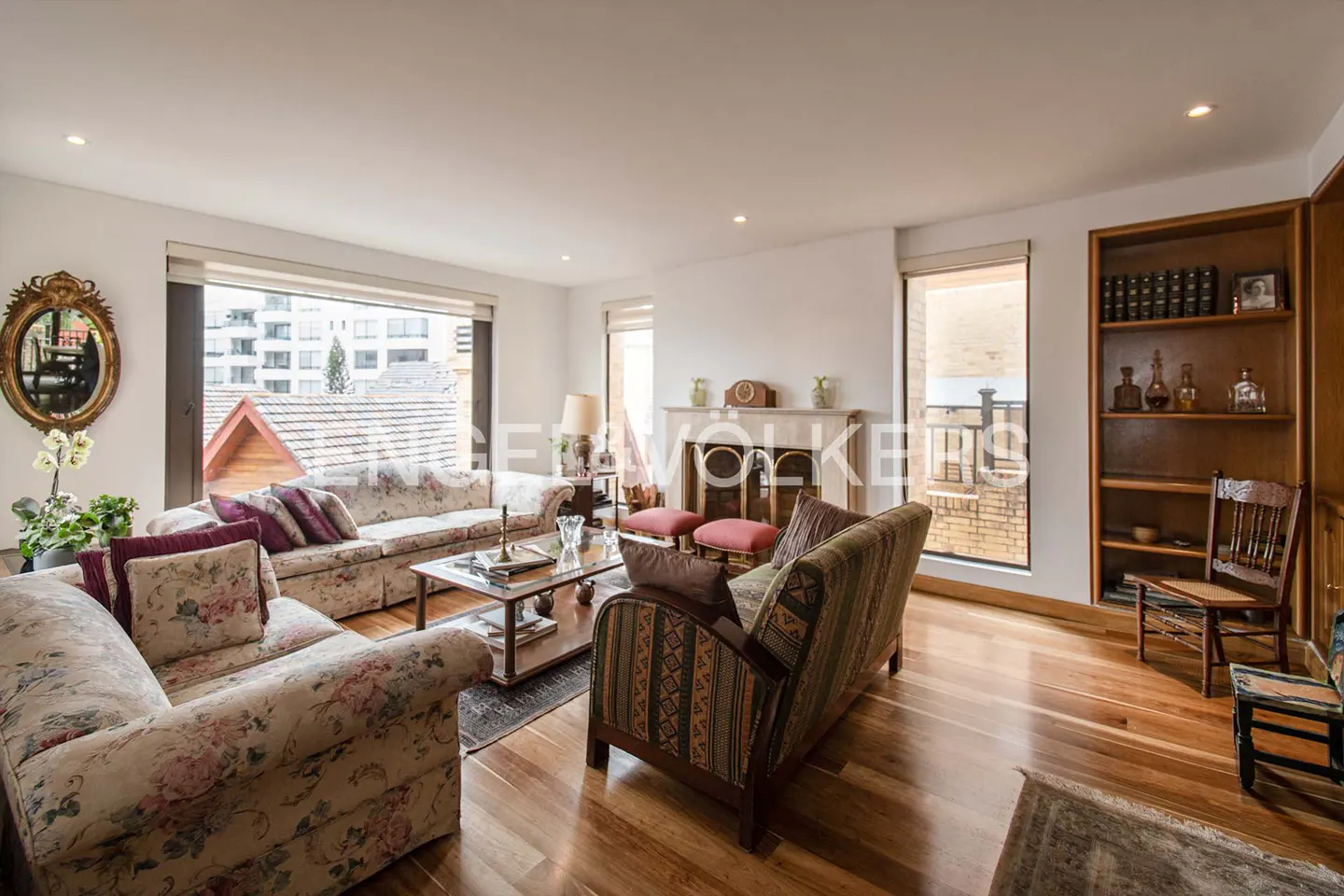 Bright living room with floral sofas, wood floors, and a fireplace. A bookshelf and large windows add to the room's charm.