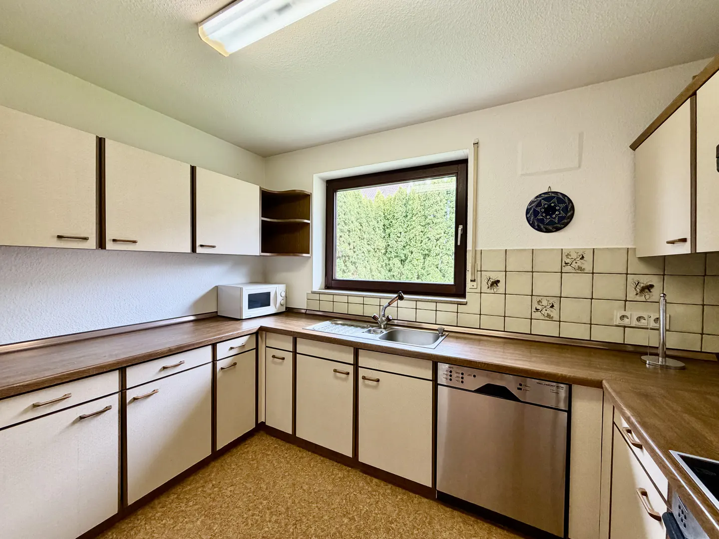 A kitchen with white cabinets, brown countertops, and a stainless steel dishwasher. A window overlooks green foliage.