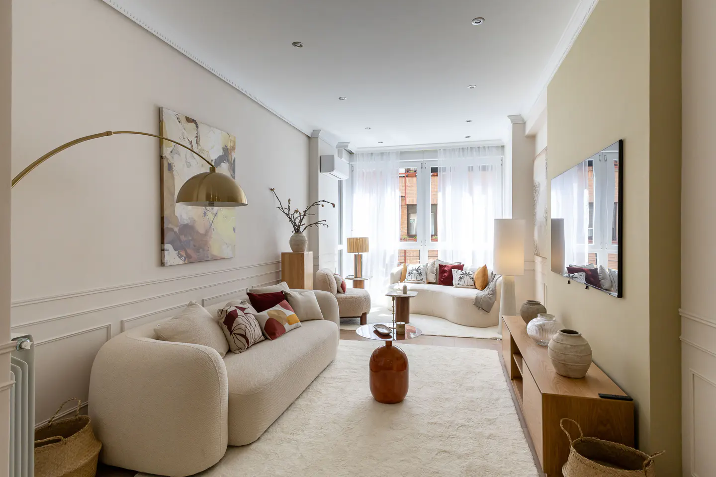 Bright living room with white walls, a large white rug, and two white sofas with colorful pillows. A gold floor lamp arches over the main sofa. A TV hangs on the wall.