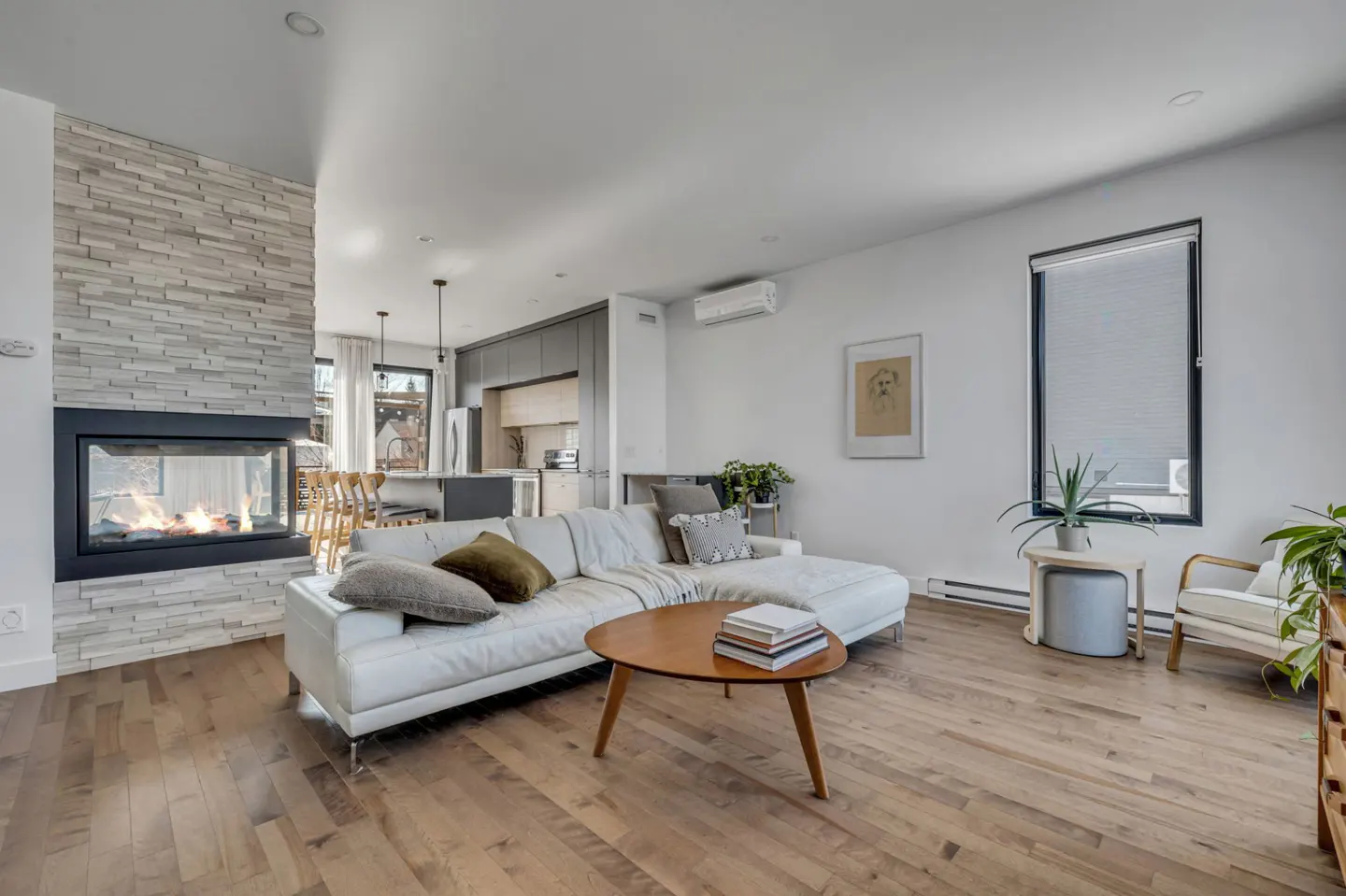 Bright, open-concept living room with hardwood floors, a white sectional sofa, and a stone fireplace. A modern kitchen is visible in the background.