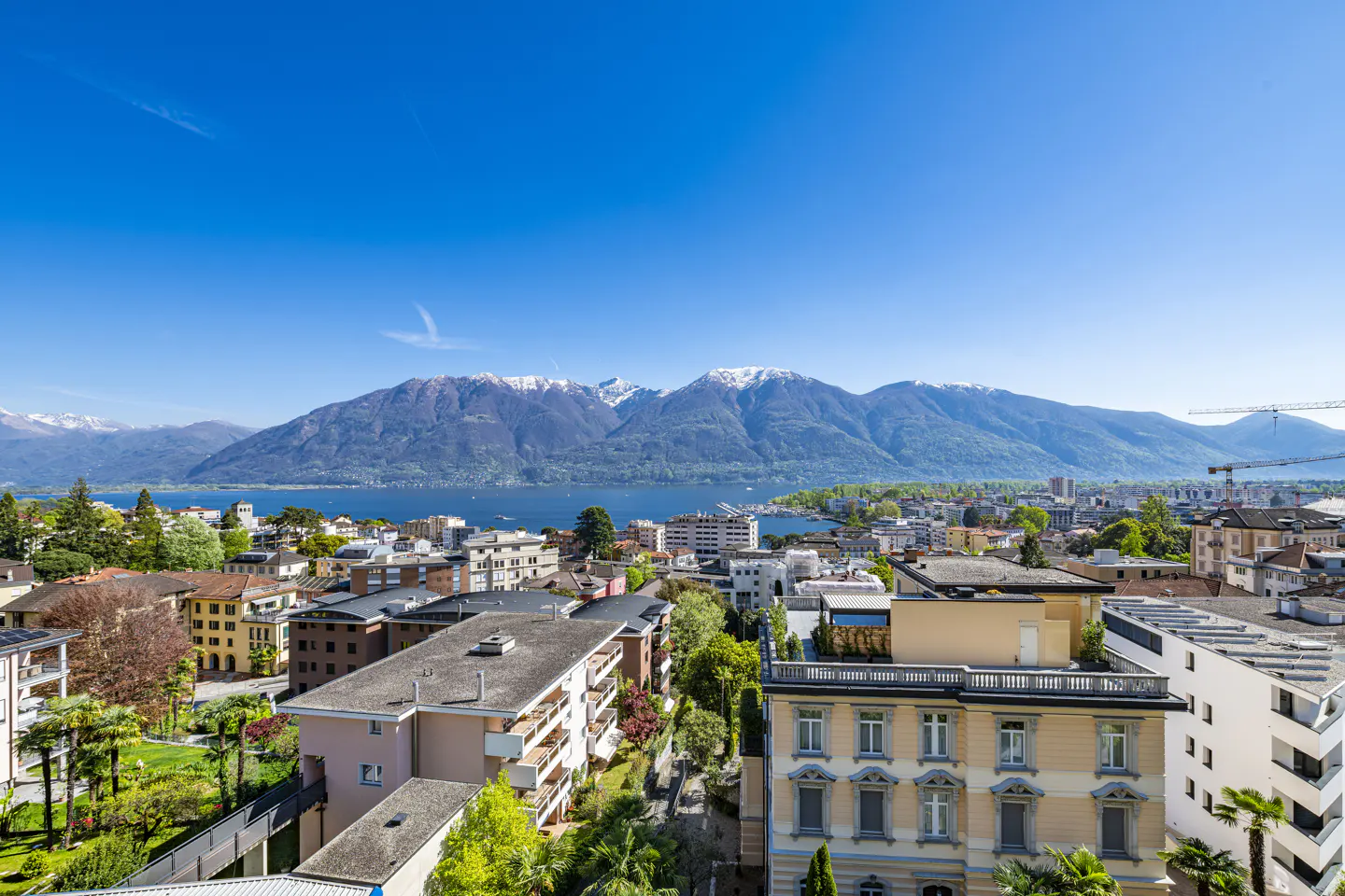 Cityscape view of Locarno, Switzerland, with buildings, Lake Maggiore, and snow-capped mountains under a clear blue sky.